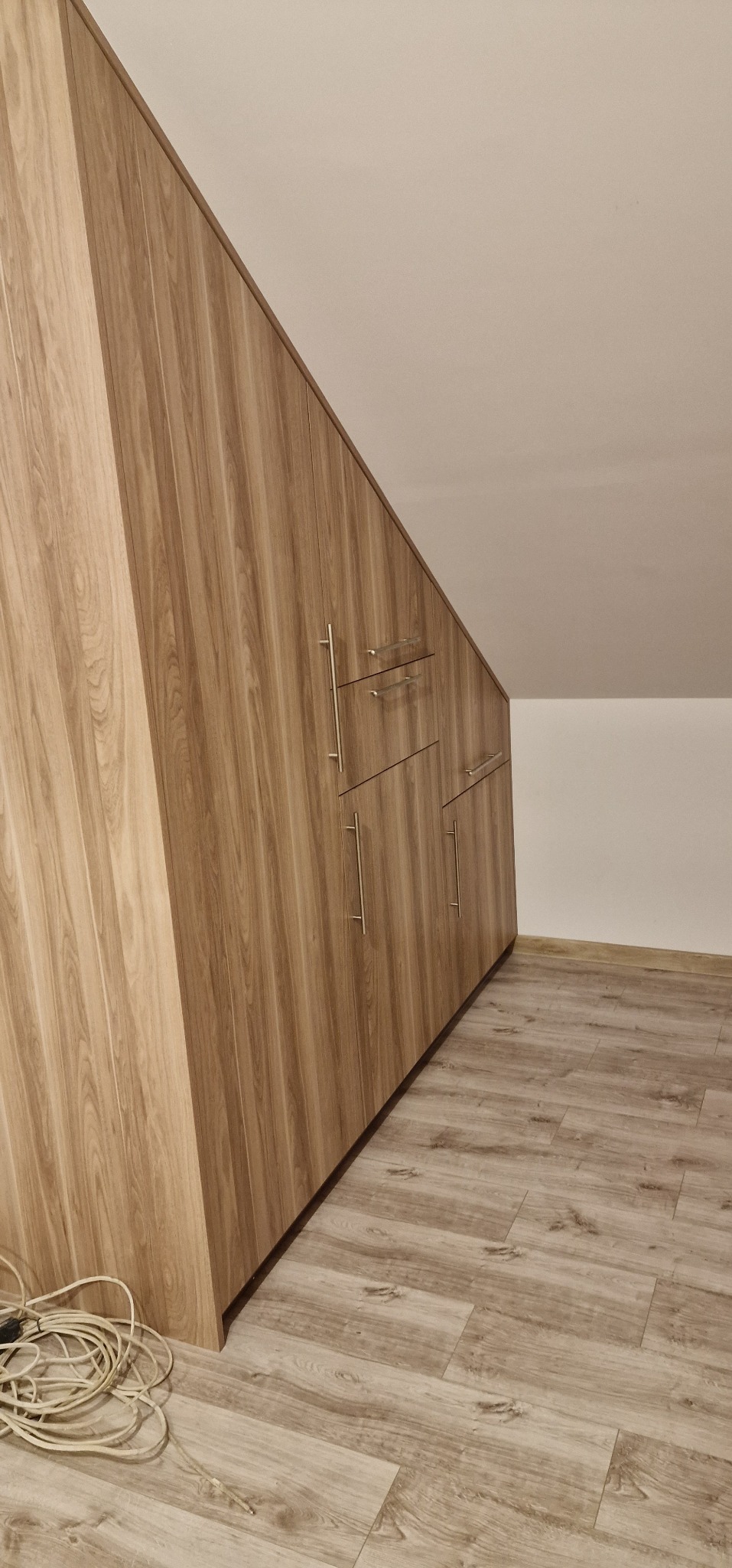 Drewniana zabudowa szafy wnękowej ze srebrnymi uchwytami, dopasowana do skosu poddasza. Na podłodze panele imitujące drewno. Widoczne kable na podłodze.