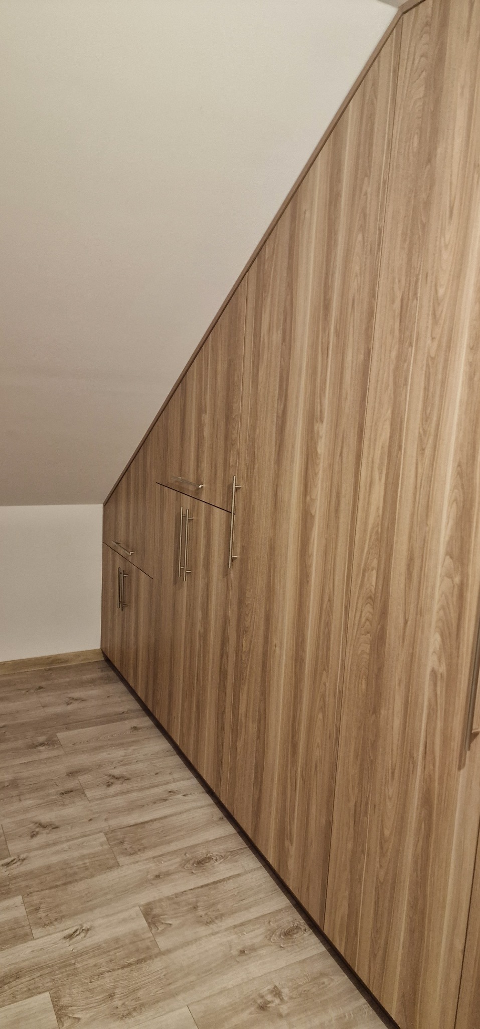 Szafa wnękowa z drewnopodobnymi frontami, dopasowana do skosu poddasza. Widoczne metalowe uchwyty i panele podłogowe w odcieniach beżu i brązu.