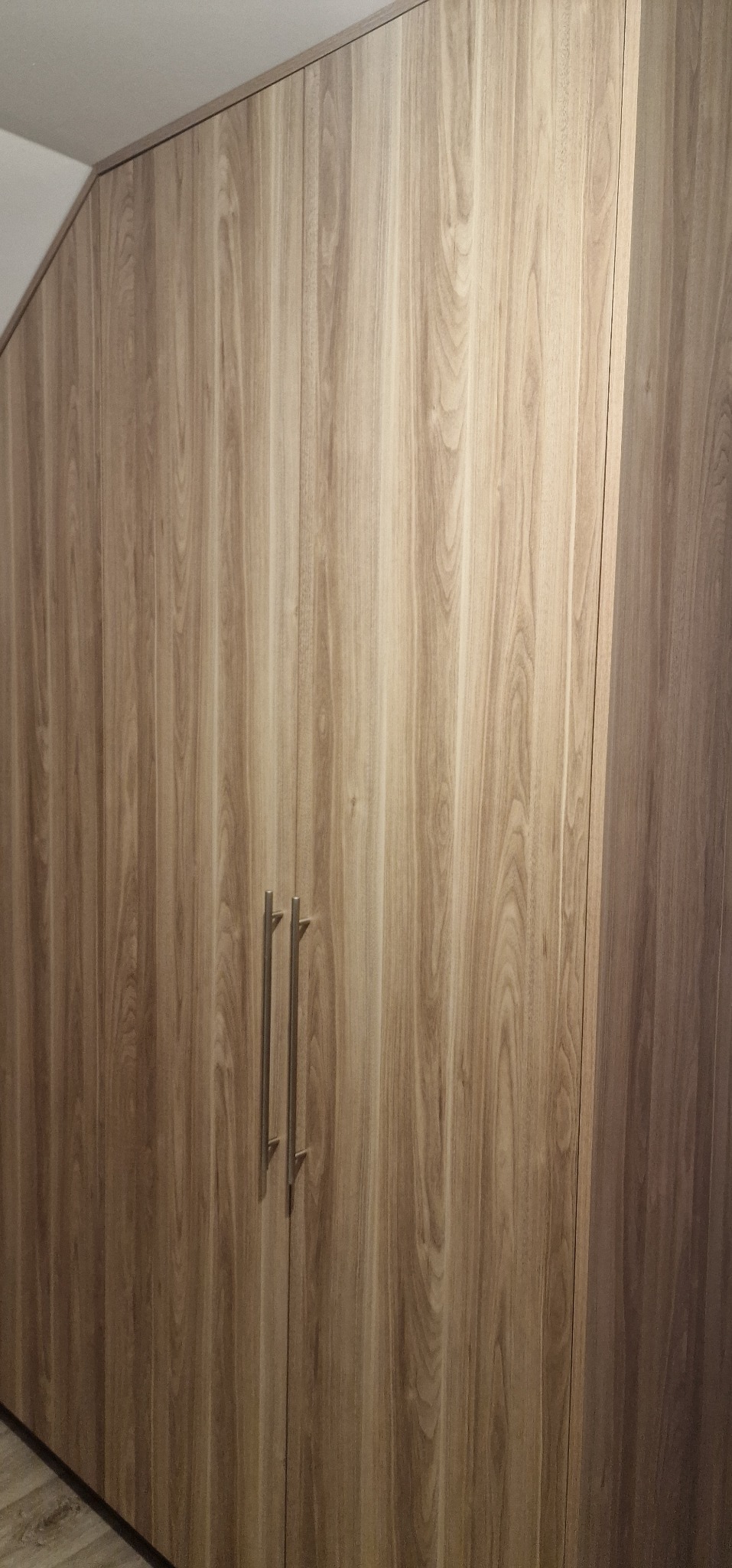 Drewniana szafa na wymiar z podłużnymi uchwytami, dopasowana do skosu sufitu, z widoczną strukturą drewna i subtelnym oświetleniem.