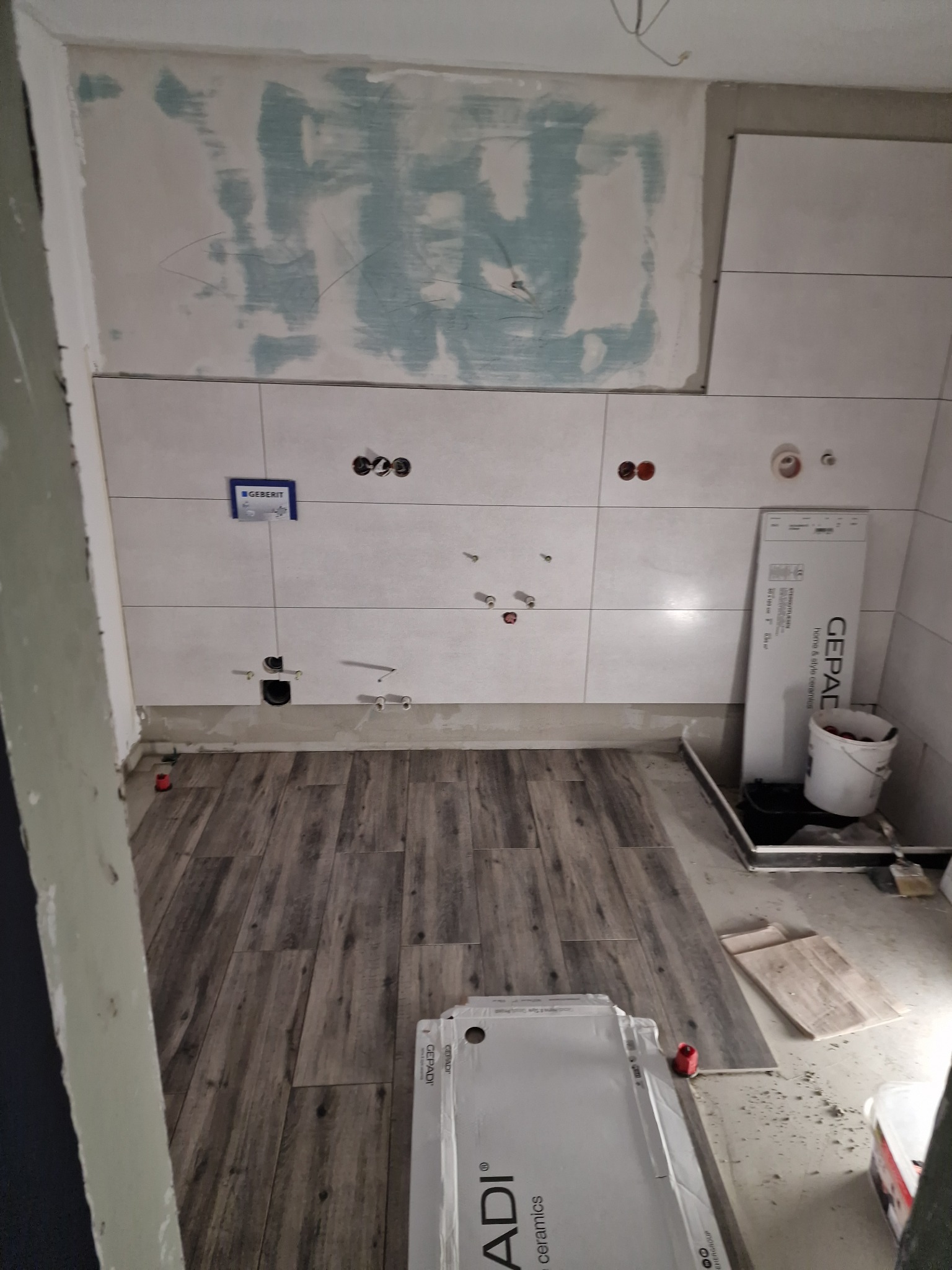 Remont łazienki w trakcie: ułożone płytki ścienne z instalacją, częściowo wykończona ściana, płytki podłogowe imitujące drewno, paczki płytek na podłodze.