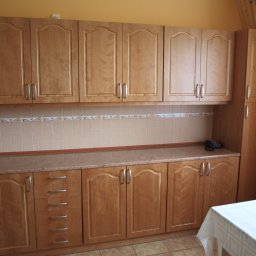 TRANSI P.U.H. - Jasne, drewniane szafki kuchenne z szufladami i blatem w Zembrzycach. Widoczne górne i dolne szafki, płytki nad blatem i fragment stołu z białym obrusem.