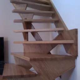 Nowoczesne, ażurowe schody drewniane z jasnego drewna. Widoczne stopnie i konstrukcja nośna, minimalistyczny design, fragment telewizora w tle.