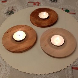 TRANSI P.U.H. - Trzy drewniane świeczniki na okrągłej serwetce z motywem świątecznym, każdy z zapaloną świeczką tealight. Widoczna agrafka na serwetce.