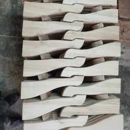 TRANSI P.U.H. - Stos drewnianych, jasnych elementów o falistym kształcie, ułożonych jeden na drugim, oddzielonych małymi klockami. Widoczne buty osoby w lewym dolnym rogu.