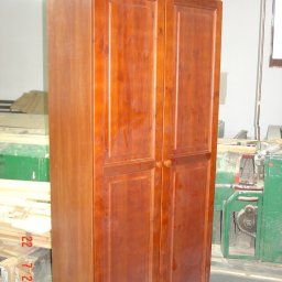 TRANSI P.U.H. - Dwudrzwiowa szafa z litego drewna w ciepłym odcieniu brązu, z wyraźnymi słojami, stoi w warsztacie stolarskim obok zielonej maszyny. Widoczne deski i karton.