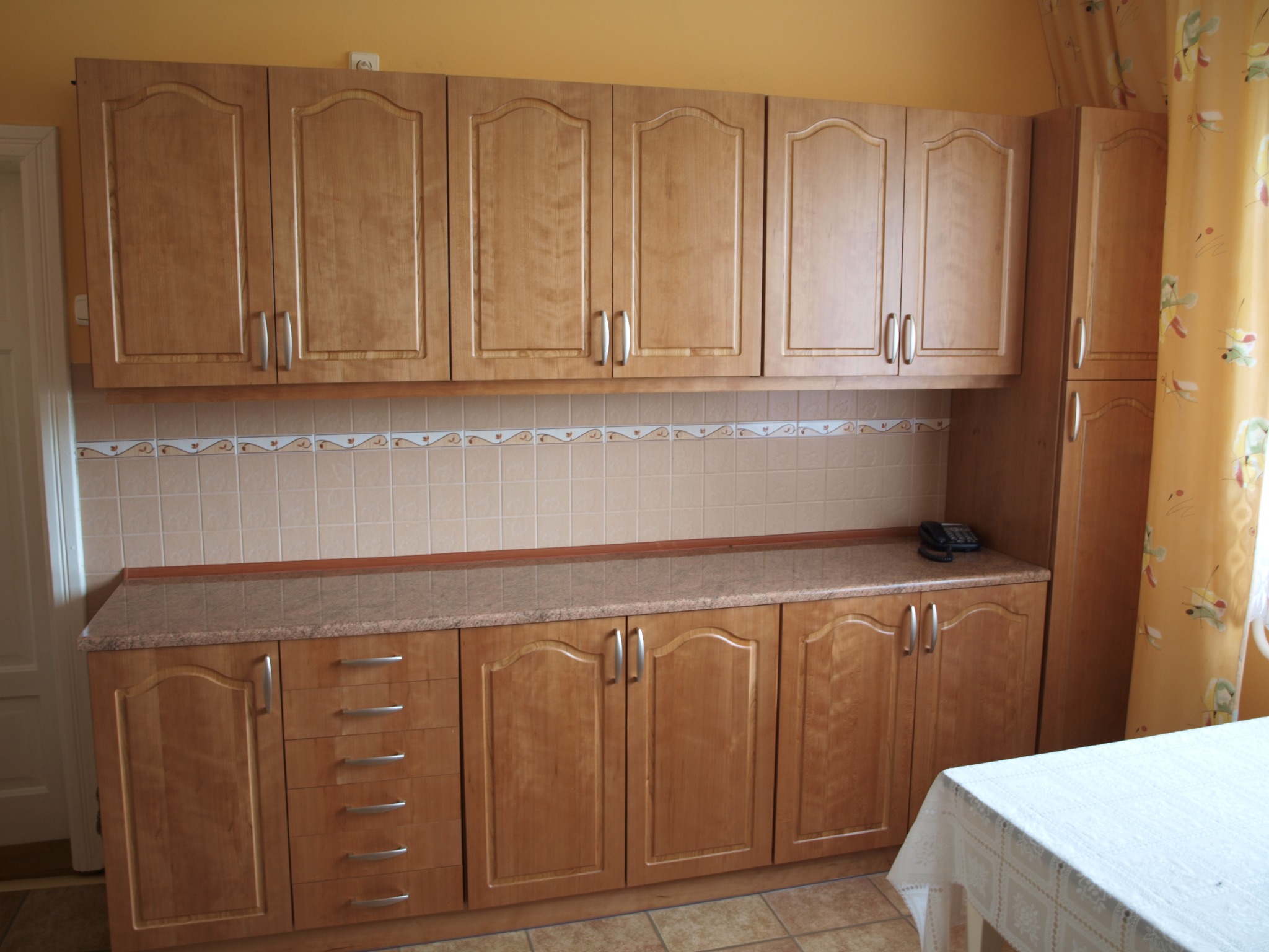 Jasne, drewniane szafki kuchenne z szufladami i blatem w Zembrzycach. Widoczne górne i dolne szafki, płytki nad blatem i fragment stołu z białym obrusem.