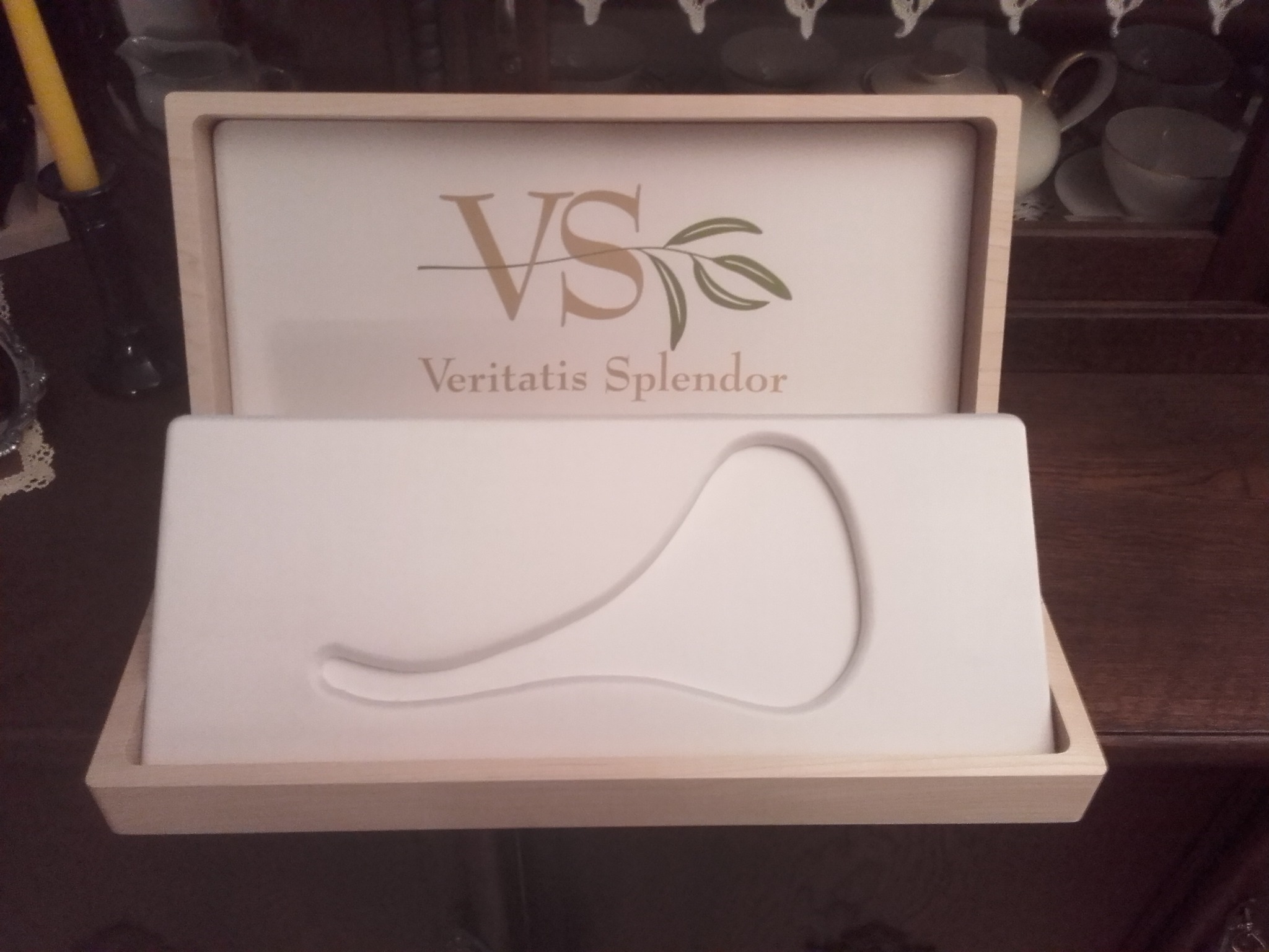 Otwarta, jasna, drewniana szkatułka z logo 'VS Veritatis Splendor' i wyprofilowanym wnętrzem na biżuterię, ustawiona na drewnianej powierzchni.