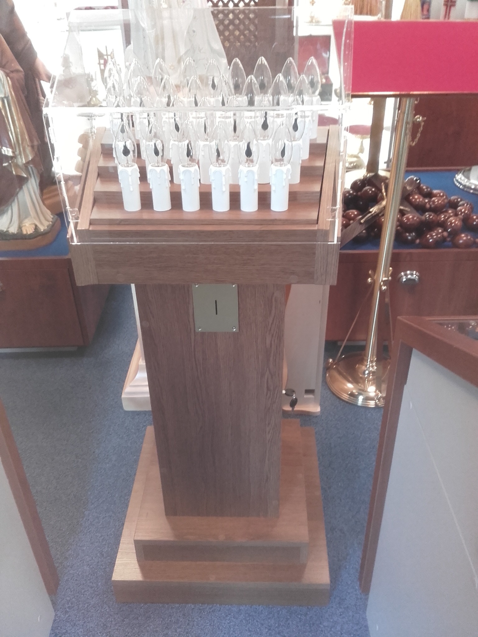 Drewniany stojak z przezroczystą obudową i rzędami elektrycznych świec w kształcie płomyków, umieszczony w kościelnym wnętrzu. Widoczna metalowa szczelina na datki.