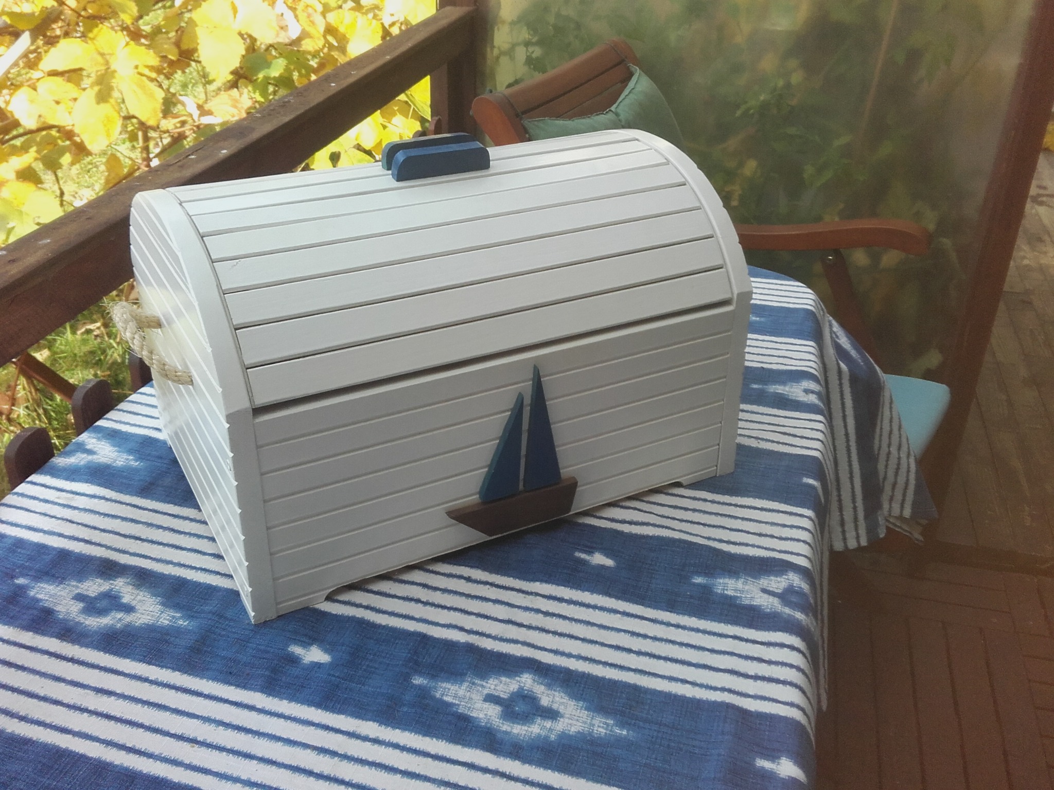 Biała, drewniana skrzynia z motywem żaglówki, na niebiesko-białym obrusie, w tle zieleń i elementy drewniane. Skrzynia dekoracyjna.