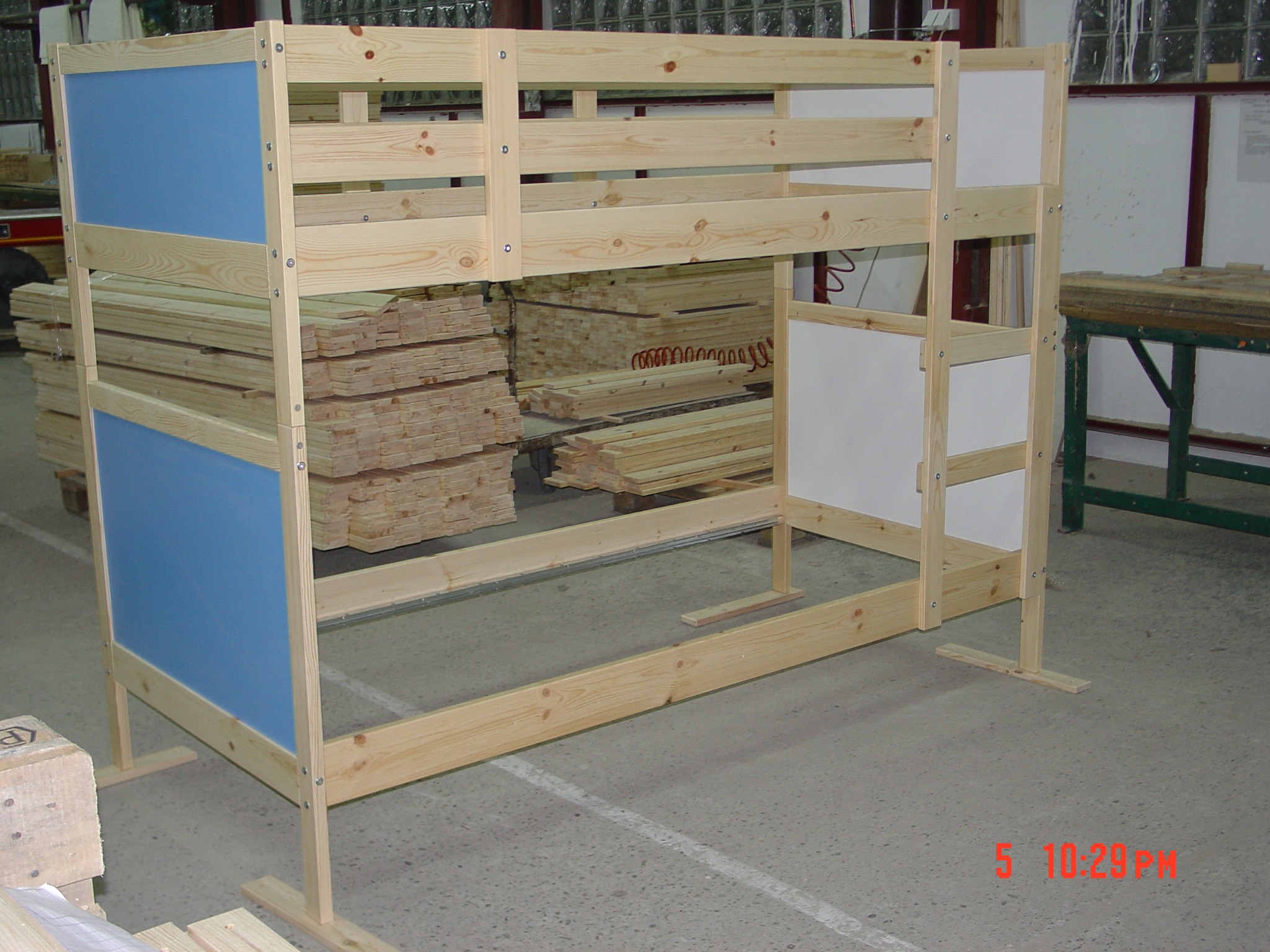 Szkielet drewnianego łóżka piętrowego z niebieskimi i białymi panelami, częściowo zmontowany w warsztacie, z zapasem drewna w tle.