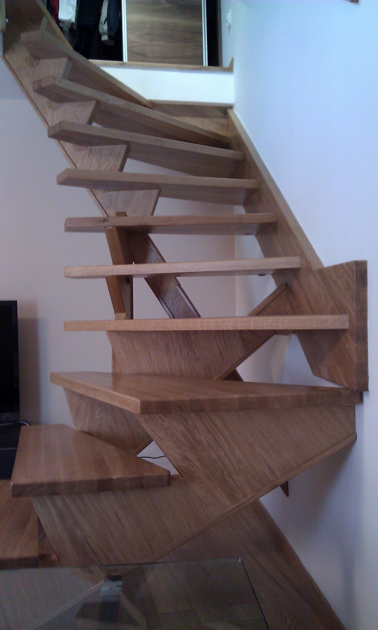 Nowoczesne, ażurowe schody drewniane z jasnego drewna. Widoczne stopnie i konstrukcja nośna, minimalistyczny design, fragment telewizora w tle.