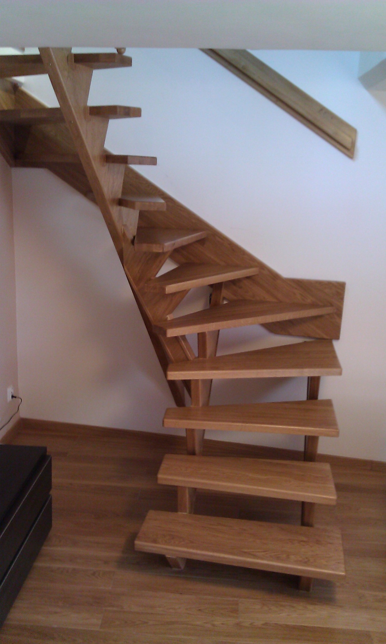 Drewniane schody o jasnym odcieniu drewna, z widoczną strukturą słojów, zamontowane w jasnym wnętrzu. Częściowo widoczna podłoga z paneli.