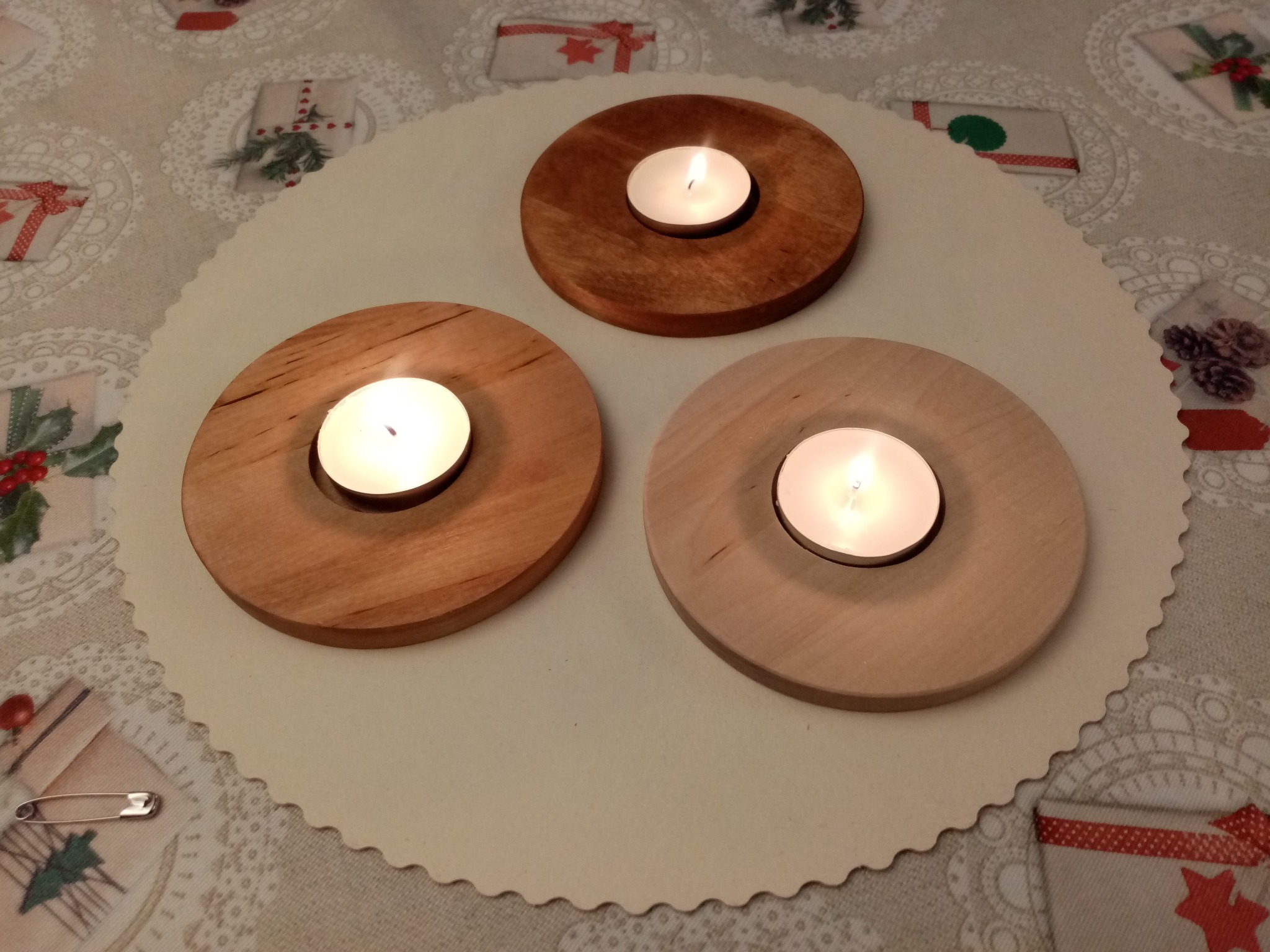 Trzy drewniane świeczniki na okrągłej serwetce z motywem świątecznym, każdy z zapaloną świeczką tealight. Widoczna agrafka na serwetce.