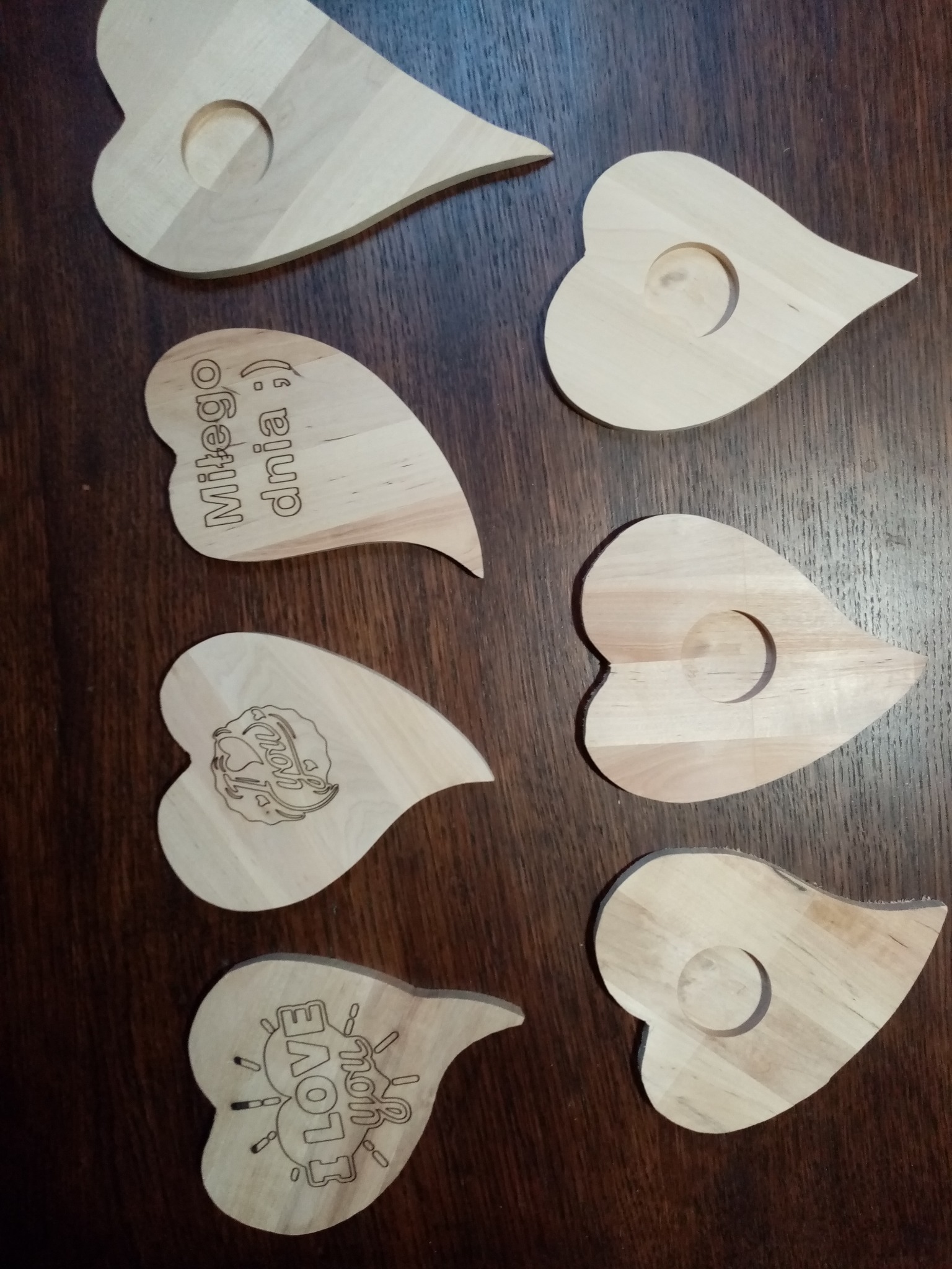 Drewniane serca z wygrawerowanymi napisami 'Miłego dnia', 'Love You' i logo, niektóre z otworem, na ciemnym, drewnianym tle. Rękodzieło.