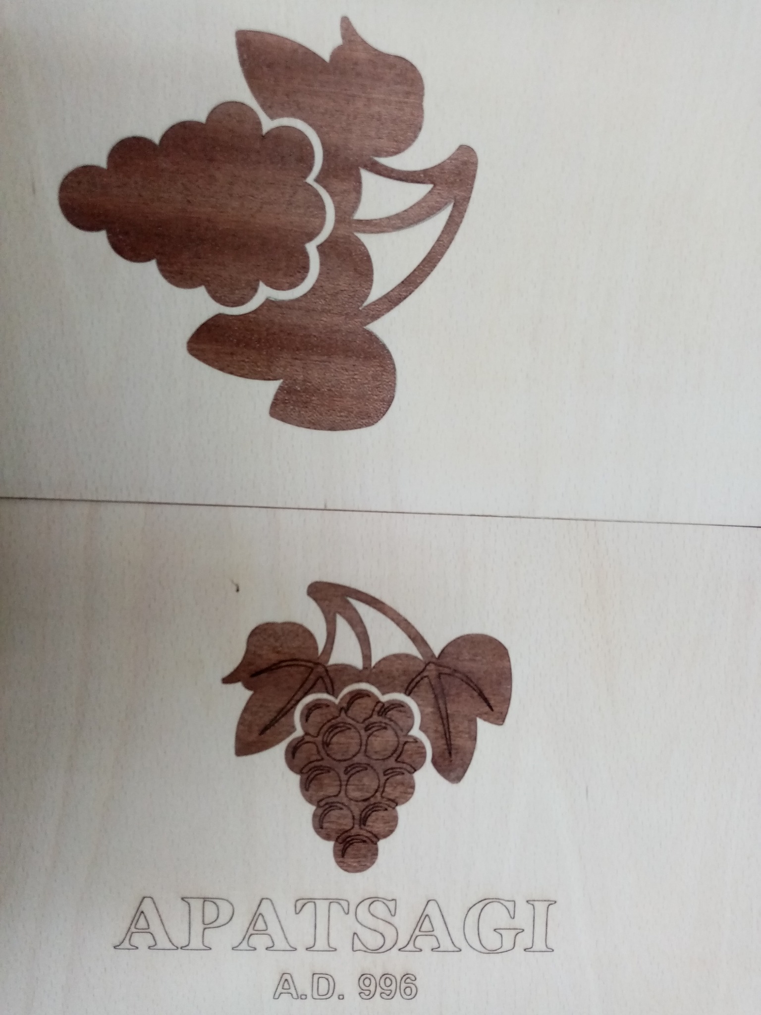 Drewniana płyta z wygrawerowanymi wzorami winogron i napisem 'APATSAGI A.D. 996'. Jasne tło, brązowe motywy. Precyzyjne wykonanie, widoczne detale.