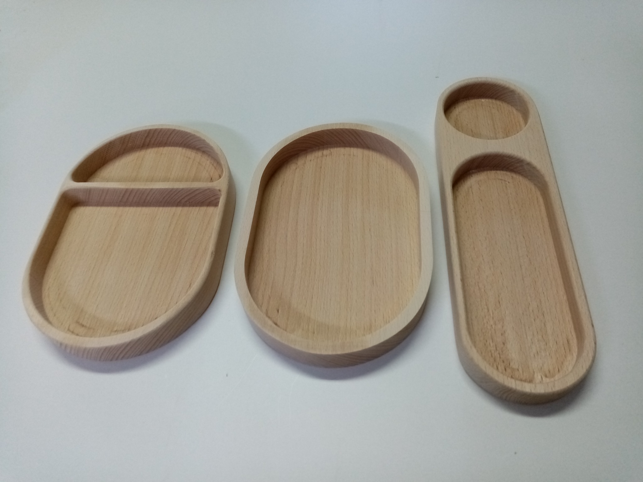 Trzy drewniane, surowe tace o owalnych kształtach, zróżnicowane pod względem podziału wewnętrznego, leżące na białym tle. Widoczny naturalny odcień drewna.