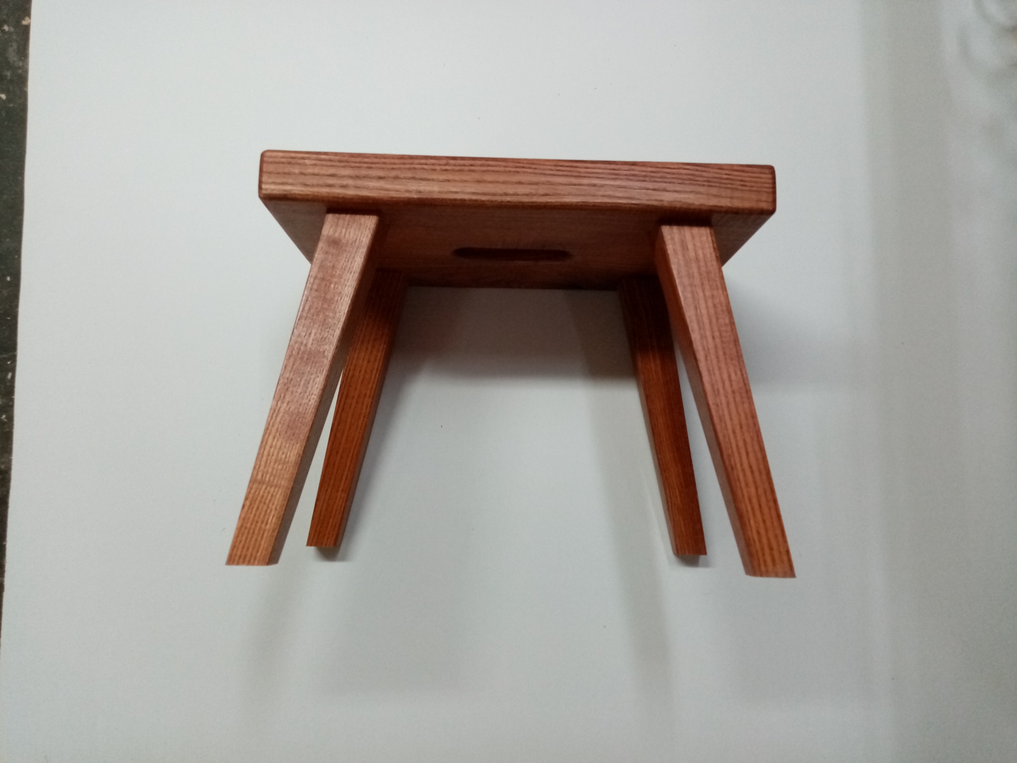 Drewniany taboret z otworem w siedzisku, wykonany z jasnego drewna, ustawiony na białym tle. Widoczne proste, skośne nogi i naturalny rysunek słojów drewna.