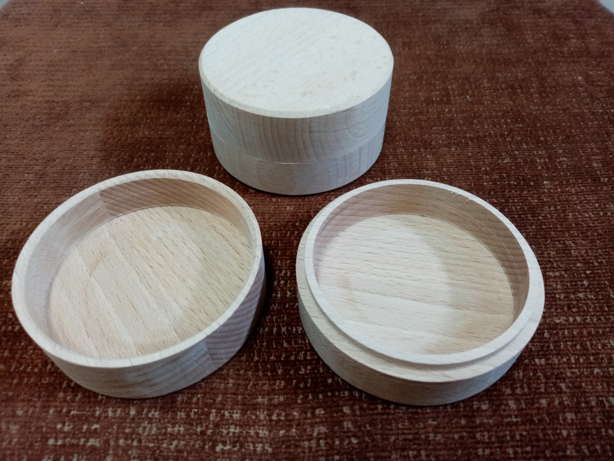 Drewniane, okrągłe pudełka z jasnego drewna na brązowym tle, z jednym pudełkiem zamkniętym i dwoma otwartymi, eksponującymi wnętrze.