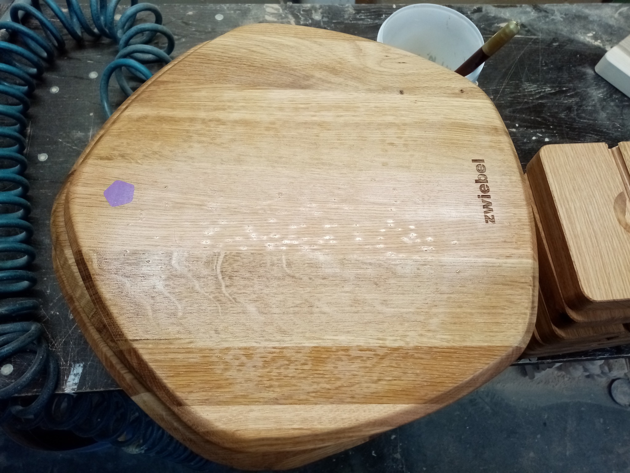 Drewniany blat z wygrawerowanym napisem 'Zwiebel', leżący na warsztacie. Widoczny fragment niebieskiego węża pneumatycznego i pojemnika z pędzlem.