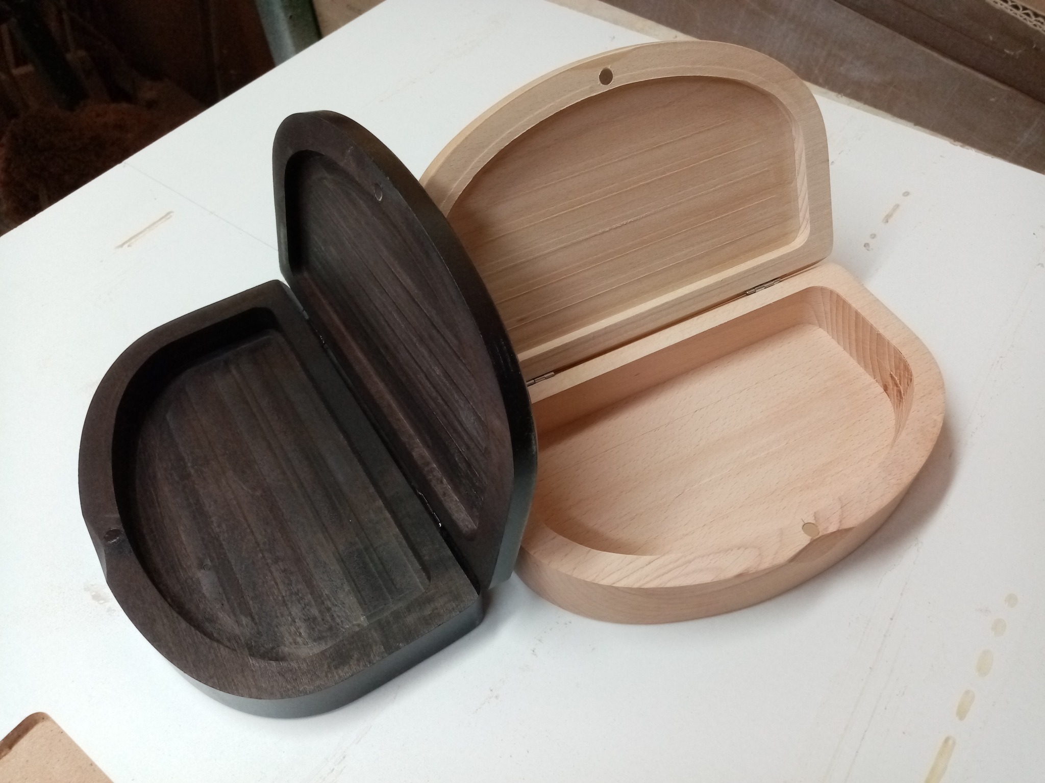 Dwie otwarte, drewniane szkatułki o zaokrąglonym kształcie: jedna z jasnego drewna, druga z ciemnego, leżące na białym blacie. Widoczne zawiasy i faktura drewna.