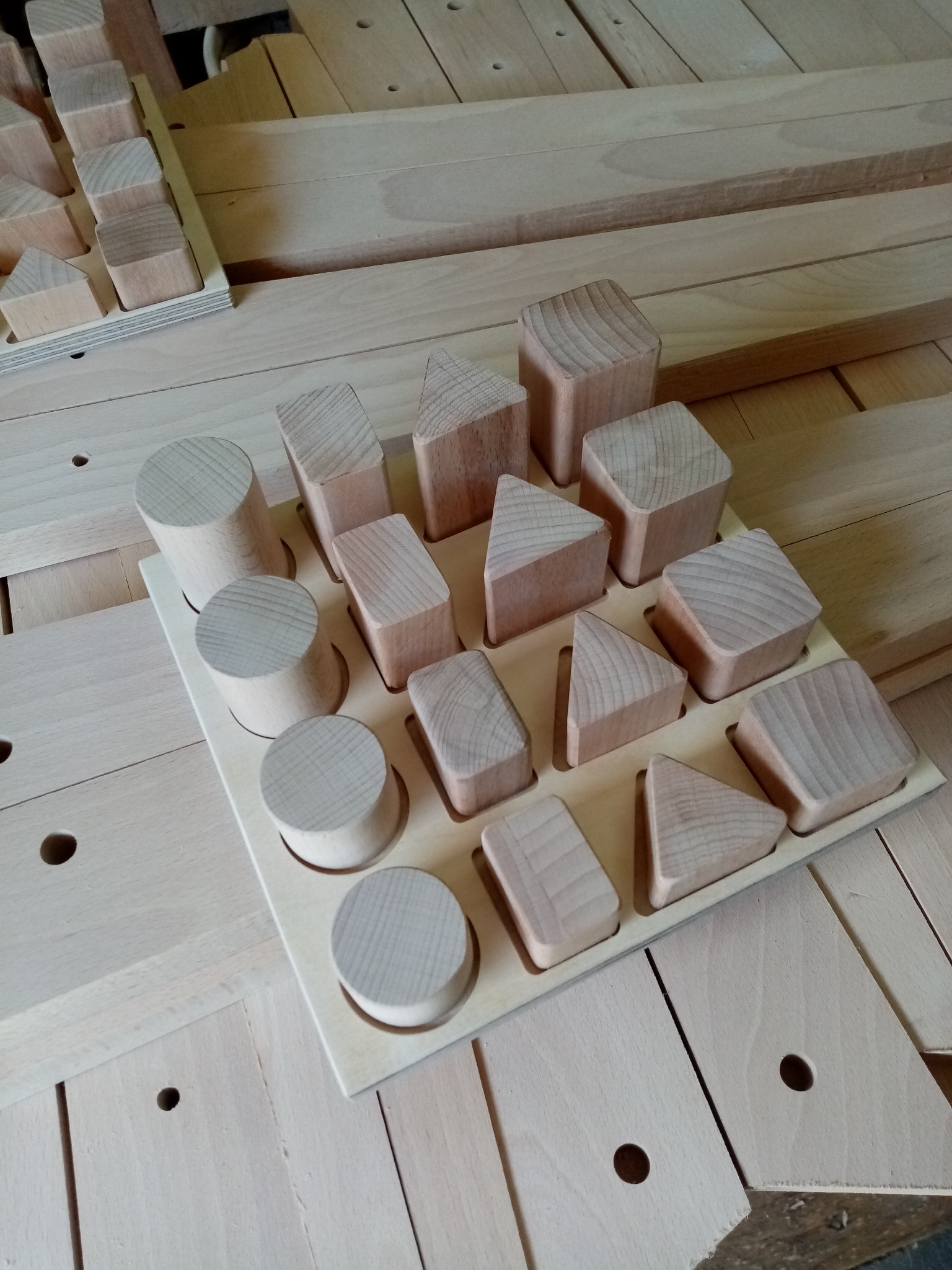 Drewniane klocki edukacyjne o różnych kształtach (koła, trójkąty, kwadraty) umieszczone w dopasowanej, jasnej, drewnianej tacy z otworami.