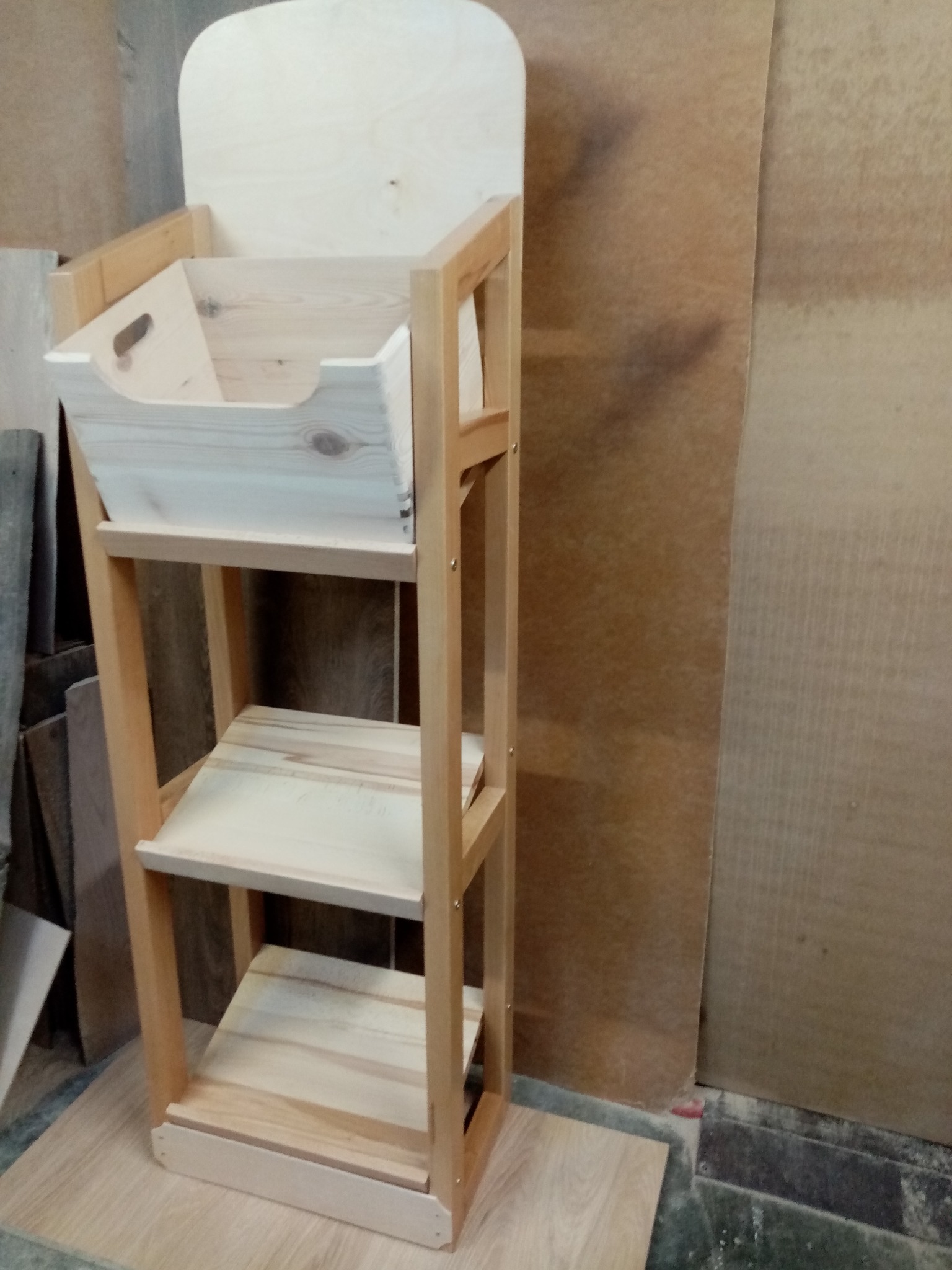 Drewniany regał z półkami i pojemnikiem, surowe drewno, jasne oświetlenie w warsztacie. Widoczne słoje drewna i detale konstrukcyjne.