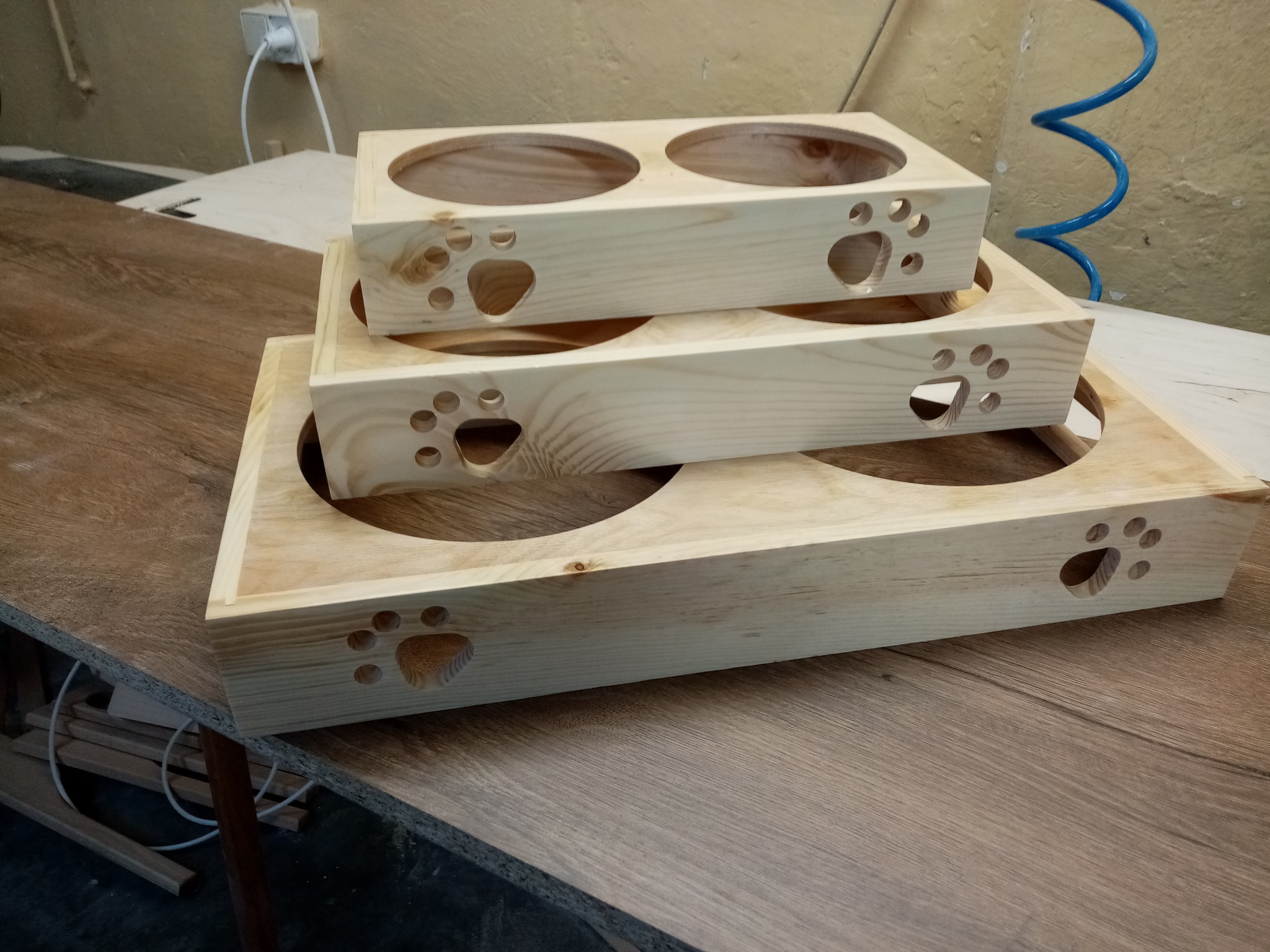 Drewniane, surowe podstawki na miski dla zwierząt, ułożone jedna na drugiej. Widoczne wycięte wzory łapek. Naturalne drewno, prosty design.