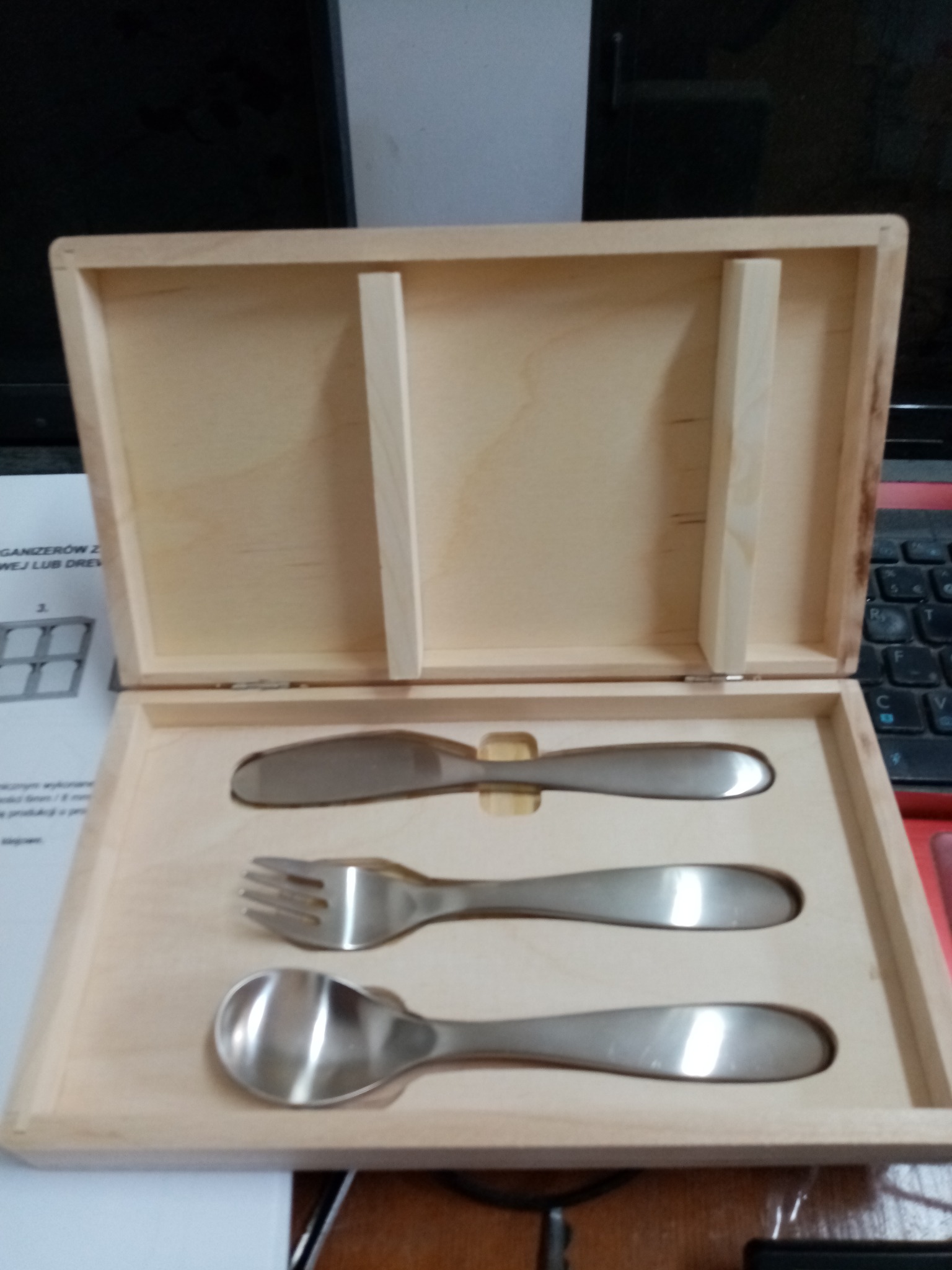 Drewniane pudełko z wyprofilowanym wnętrzem na sztućce: nóż, widelec i łyżka. Precyzyjna obróbka CNC drewna. Światło odbija się od metalowych sztućców.