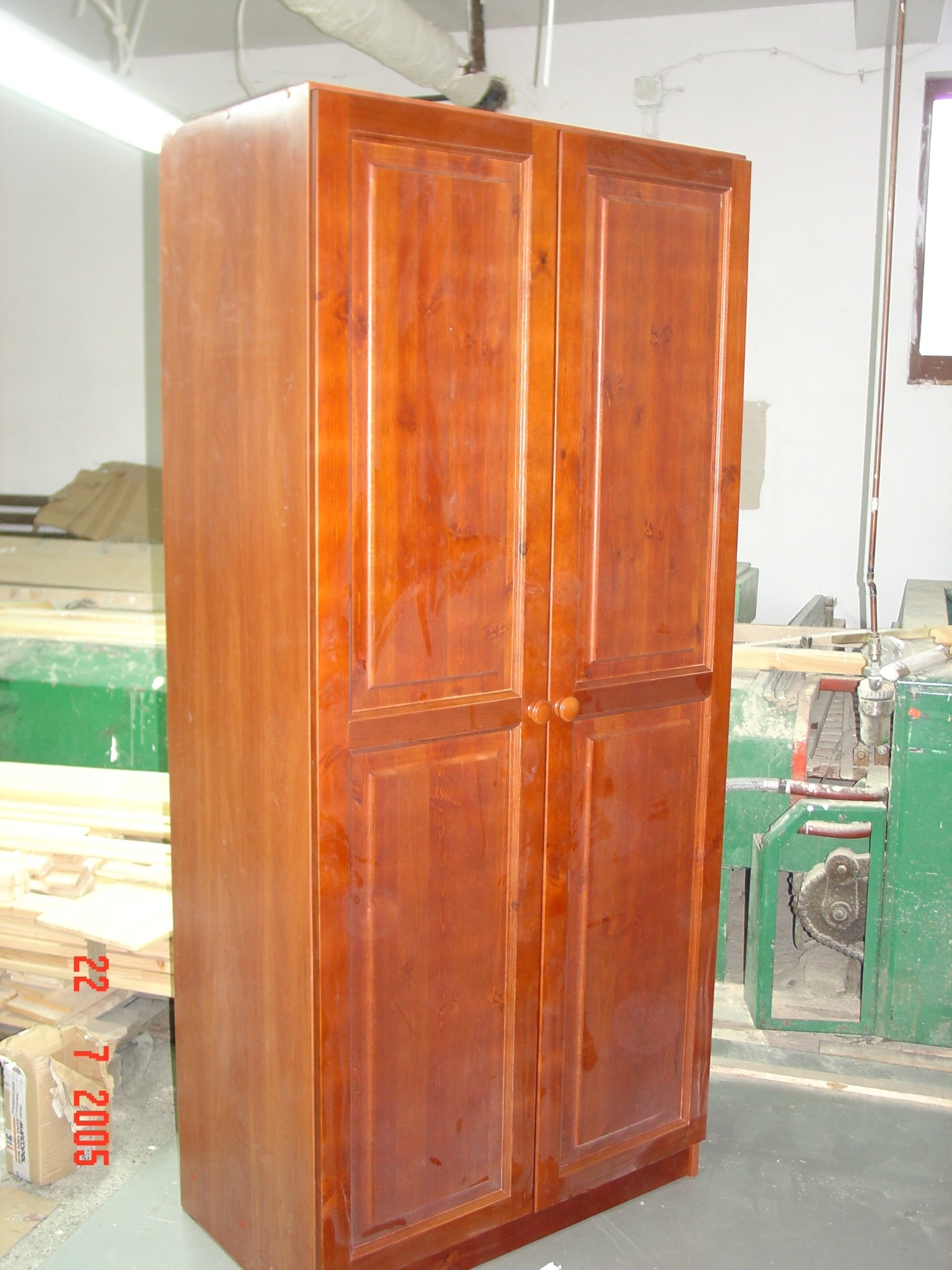 Dwudrzwiowa szafa z litego drewna w ciepłym odcieniu brązu, z wyraźnymi słojami, stoi w warsztacie stolarskim obok zielonej maszyny. Widoczne deski i karton.