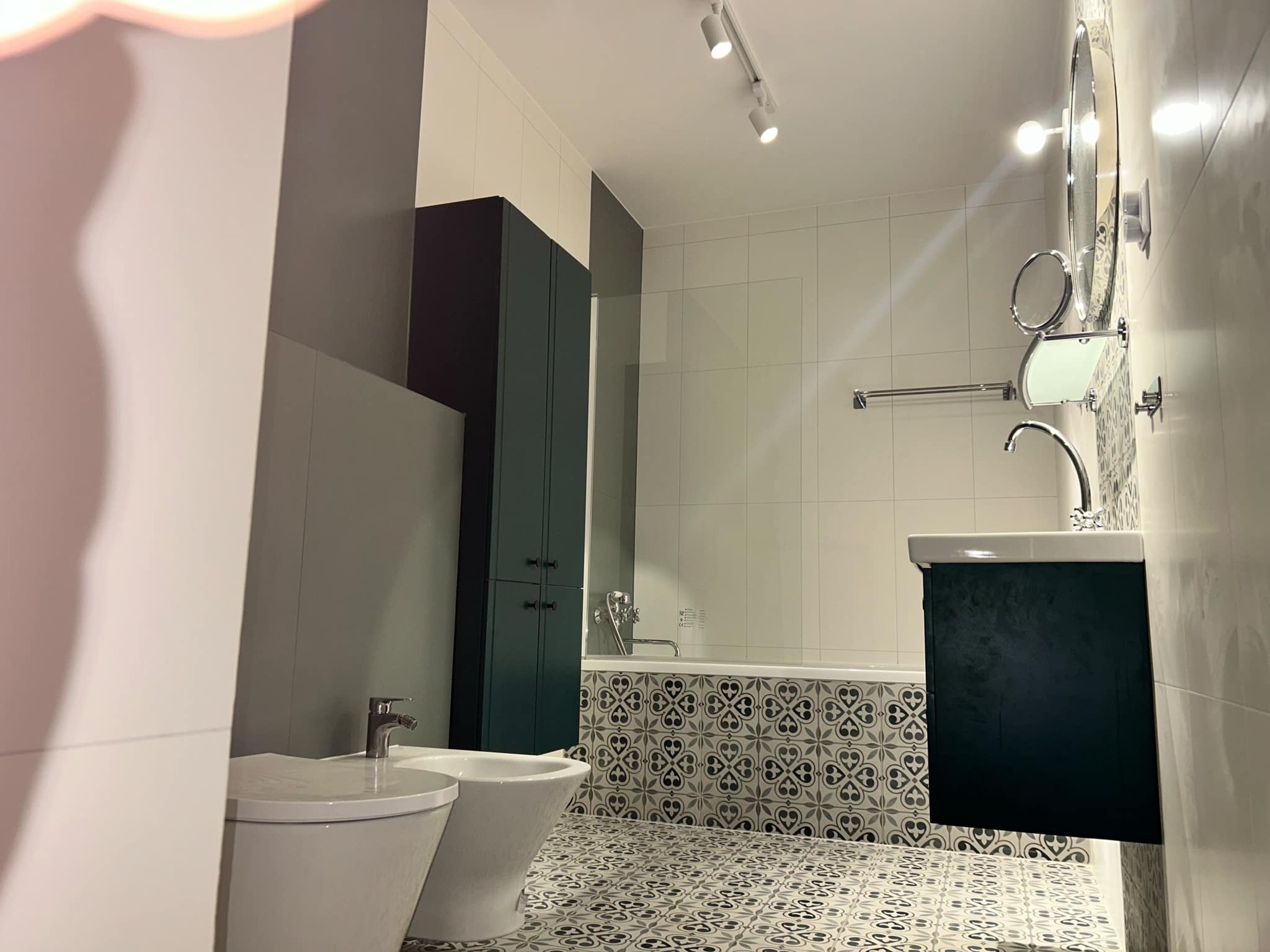 Nowoczesna łazienka w odcieniach szarości i bieli z akcentami ciemnej zieleni. Uwagę przykuwają wzorzyste płytki podłogowe i obudowa wanny.