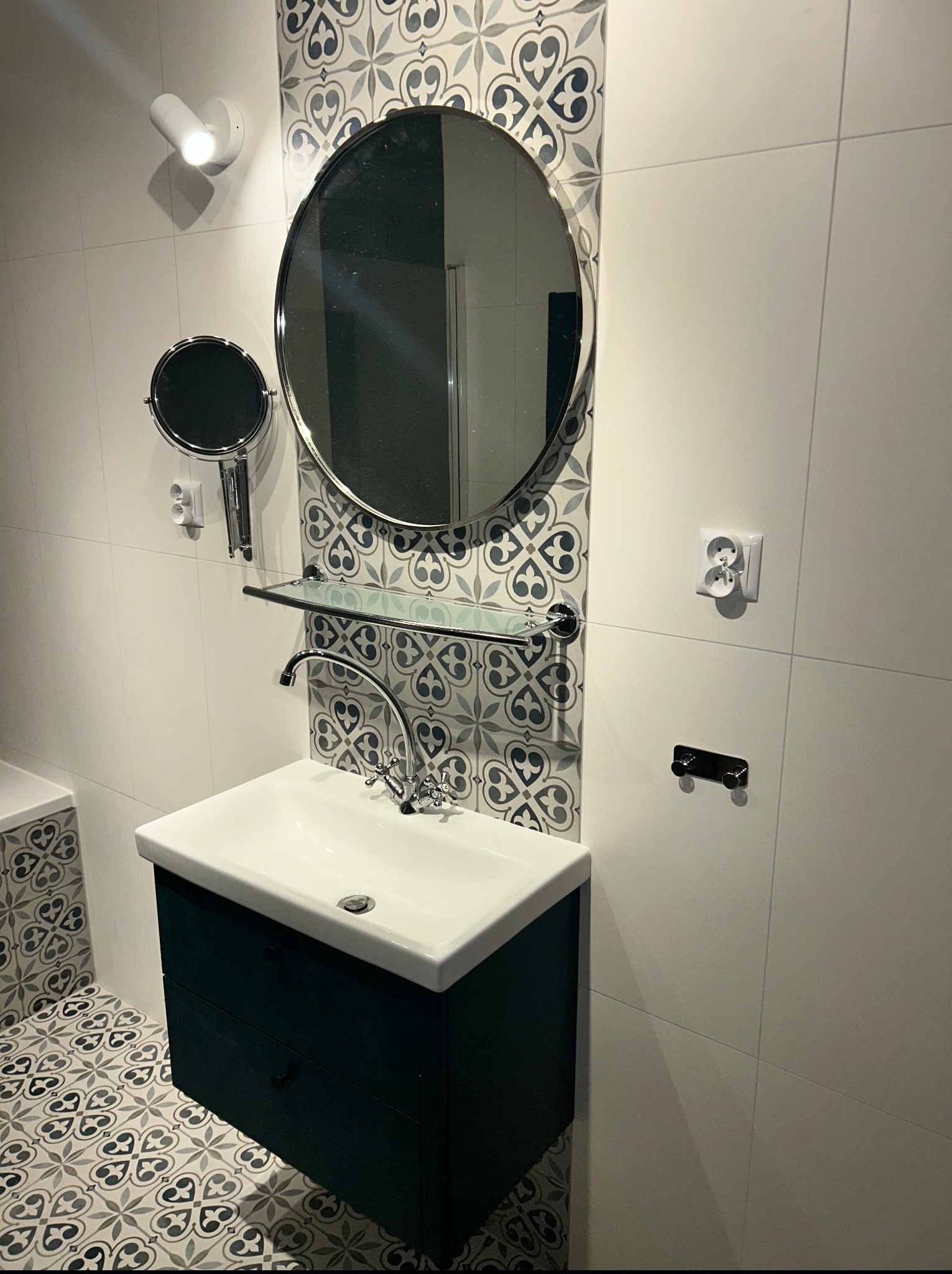 Wykończona łazienka z umywalką w szafce, okrągłym lustrem i ozdobnymi płytkami w stylu retro. Widoczny fragment wanny i nowoczesne oświetlenie.