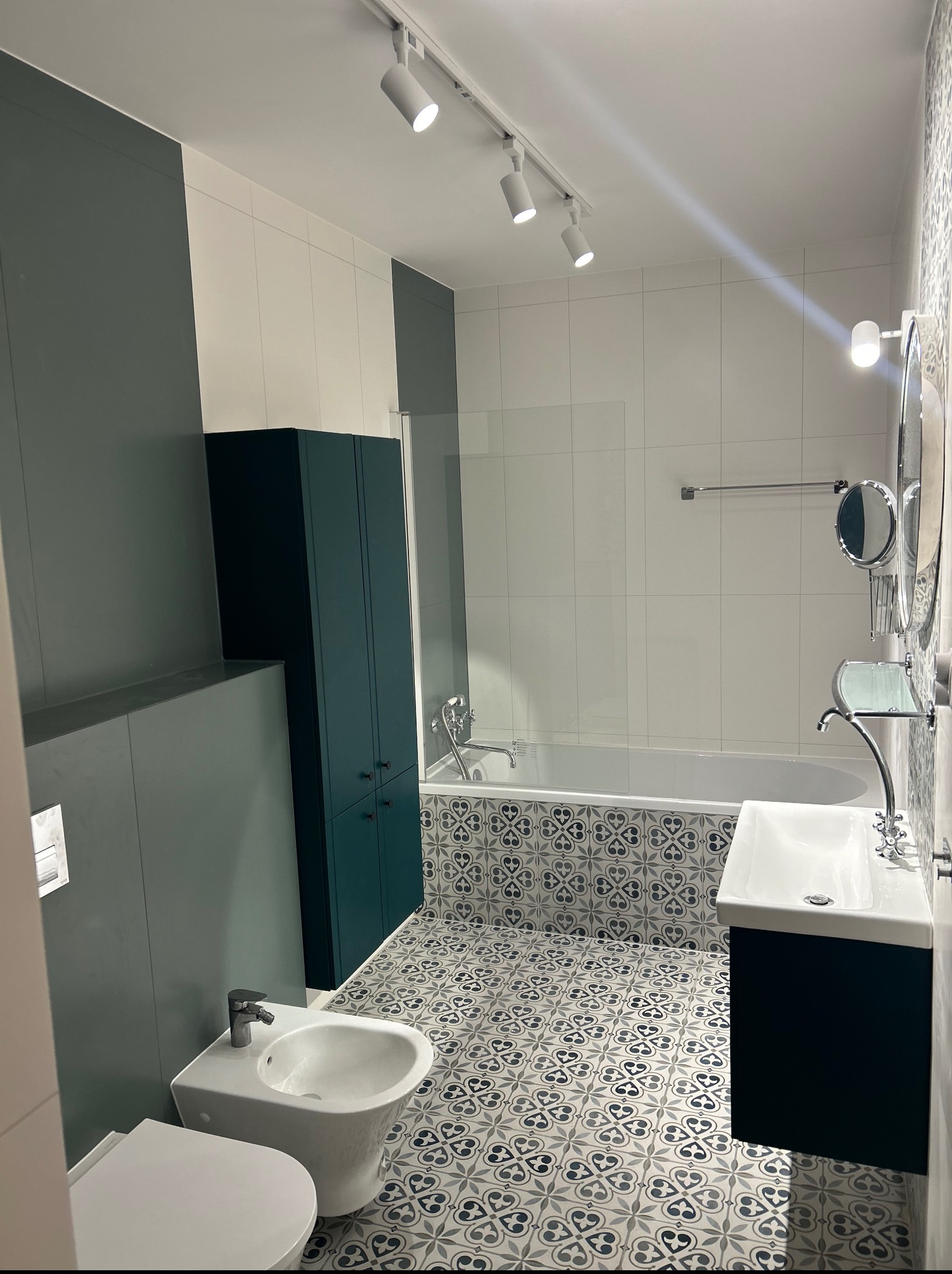 Nowoczesna łazienka po remoncie: białe płytki, wzorzysta podłoga, zielona szafka i oświetlenie punktowe. Minimalistyczny design i funkcjonalne wyposażenie.