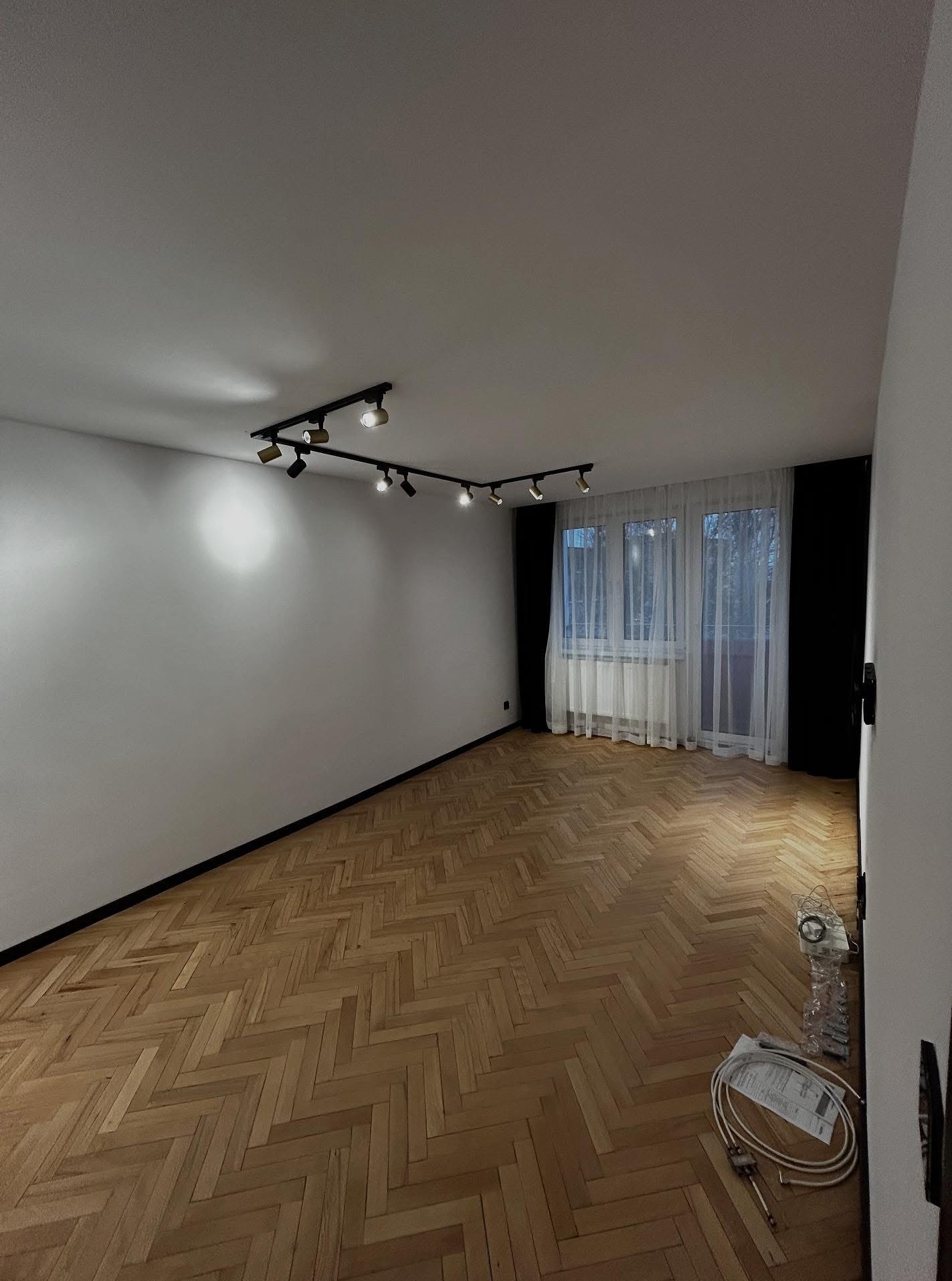 Wykończone wnętrze pokoju z parkietem w jodełkę, białymi ścianami i nowoczesnym oświetleniem na suficie. Widoczne okno z firankami i czarne zasłony.