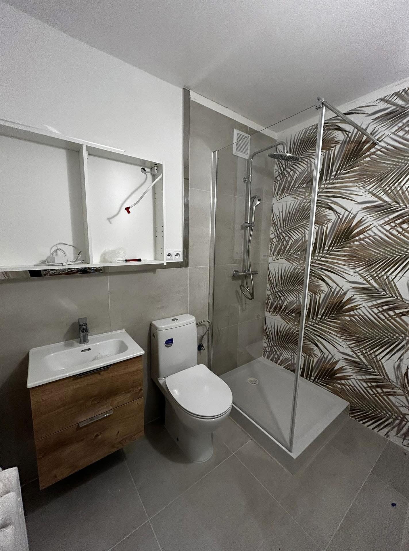 Wykończona łazienka z kabiną prysznicową typu walk-in, umywalką z drewnianą szafką i toaletą. Na ścianie tapeta z motywem palm. Częściowo widoczna szafka do montażu.