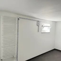 Biały grzejnik drabinkowy na ścianie, obok okno dachowe i listwy montażowe. Minimalistyczne wnętrze, czarna podłoga, świeżo po remoncie.