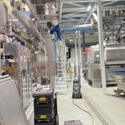 WAYCO SPÓŁKA Z OGRANICZONĄ ODPOWIEDZIALNOŚCIĄ - Pracownik w kombinezonie na drabinie, naprawia skomplikowaną maszynerię w hali produkcyjnej. Widoczne skrzynki narzędziowe i myjka ciśnieniowa. Jasne oświetlenie.