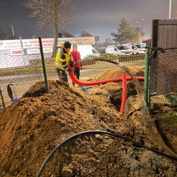 WAYCO SPÓŁKA Z OGRANICZONĄ ODPOWIEDZIALNOŚCIĄ - Prace ziemne i układanie rur w Złotoryi. Dwóch pracowników w odblaskowych ubraniach przy wykopie. W tle budynek z reklamą klimatyzacji i monitoringu.