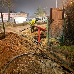 WAYCO SPÓŁKA Z OGRANICZONĄ ODPOWIEDZIALNOŚCIĄ - Prace ziemne przy układaniu kabli w ziemi. Widoczny pracownik w odblaskowej kamizelce i czerwone rury osłonowe. W tle budynek z reklamą instalacji elektrycznych.