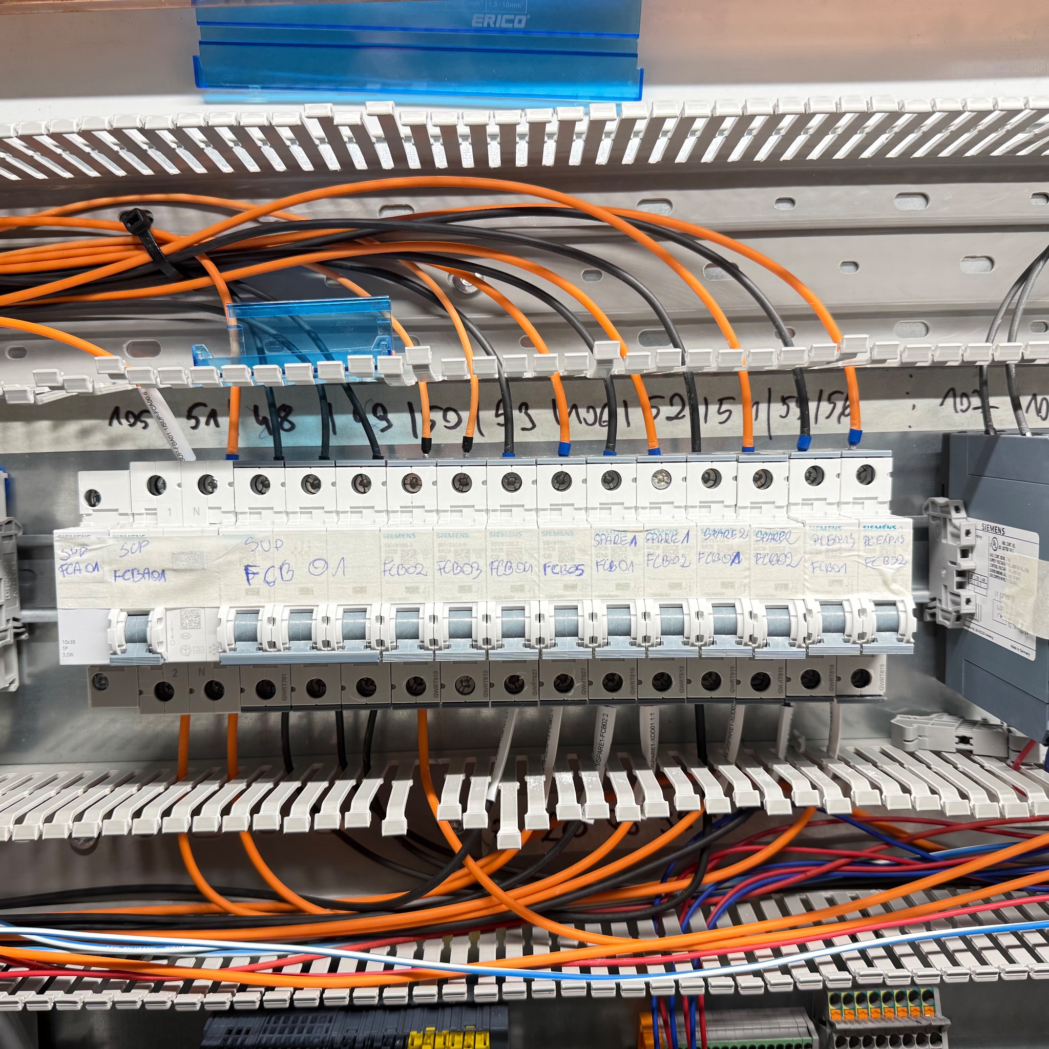 Rozdzielnica elektryczna z bezpiecznikami Siemens, okablowaniem w kolorach pomarańczowym i czarnym, oraz oznaczeniami na taśmie. Widoczne szyny montażowe i elementy sterujące.