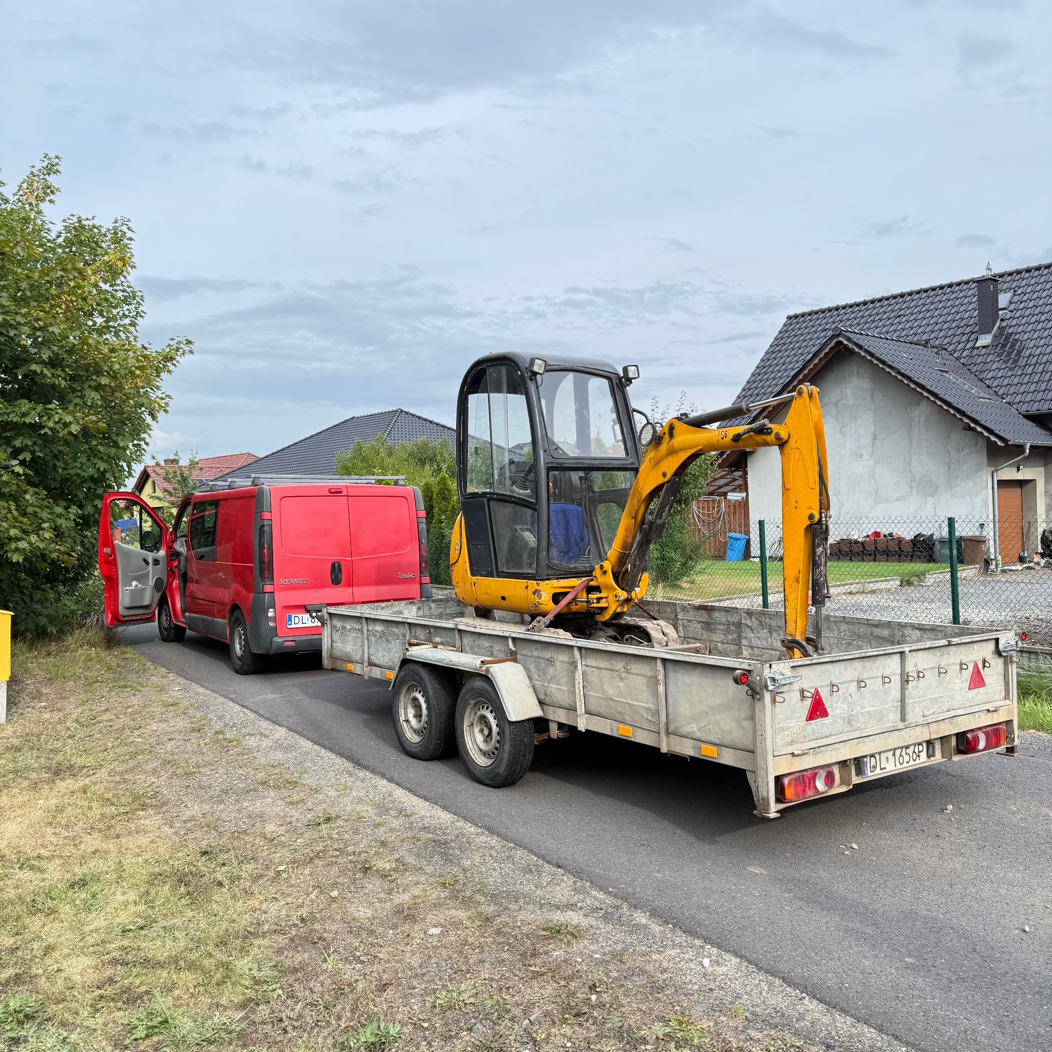 Żółta mini koparka na przyczepie za czerwonym vanem na tle domu i zieleni. Sprzęt gotowy do prac ziemnych lub wyburzeniowych.