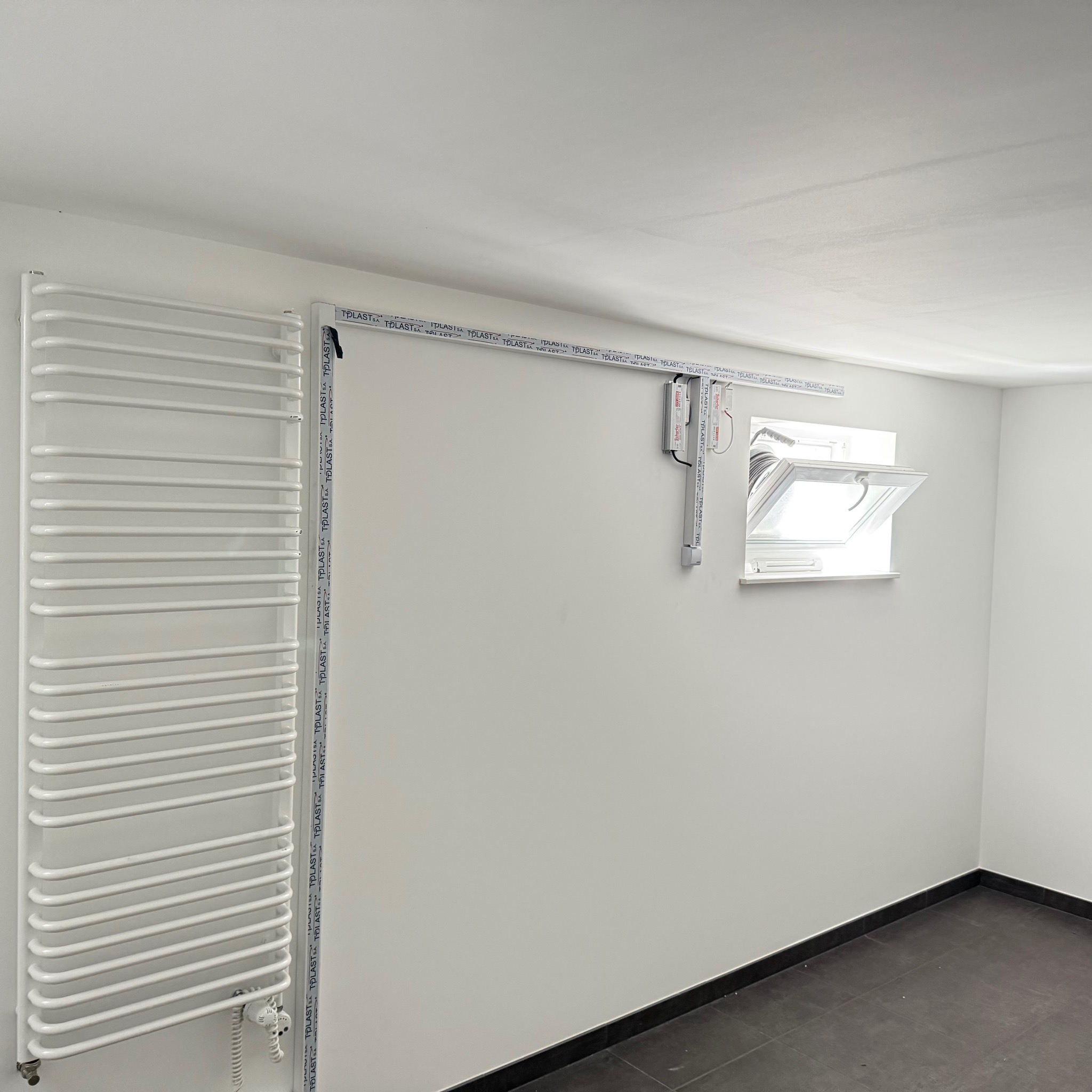 Biały grzejnik drabinkowy na ścianie, obok okno dachowe i listwy montażowe. Minimalistyczne wnętrze, czarna podłoga, świeżo po remoncie.