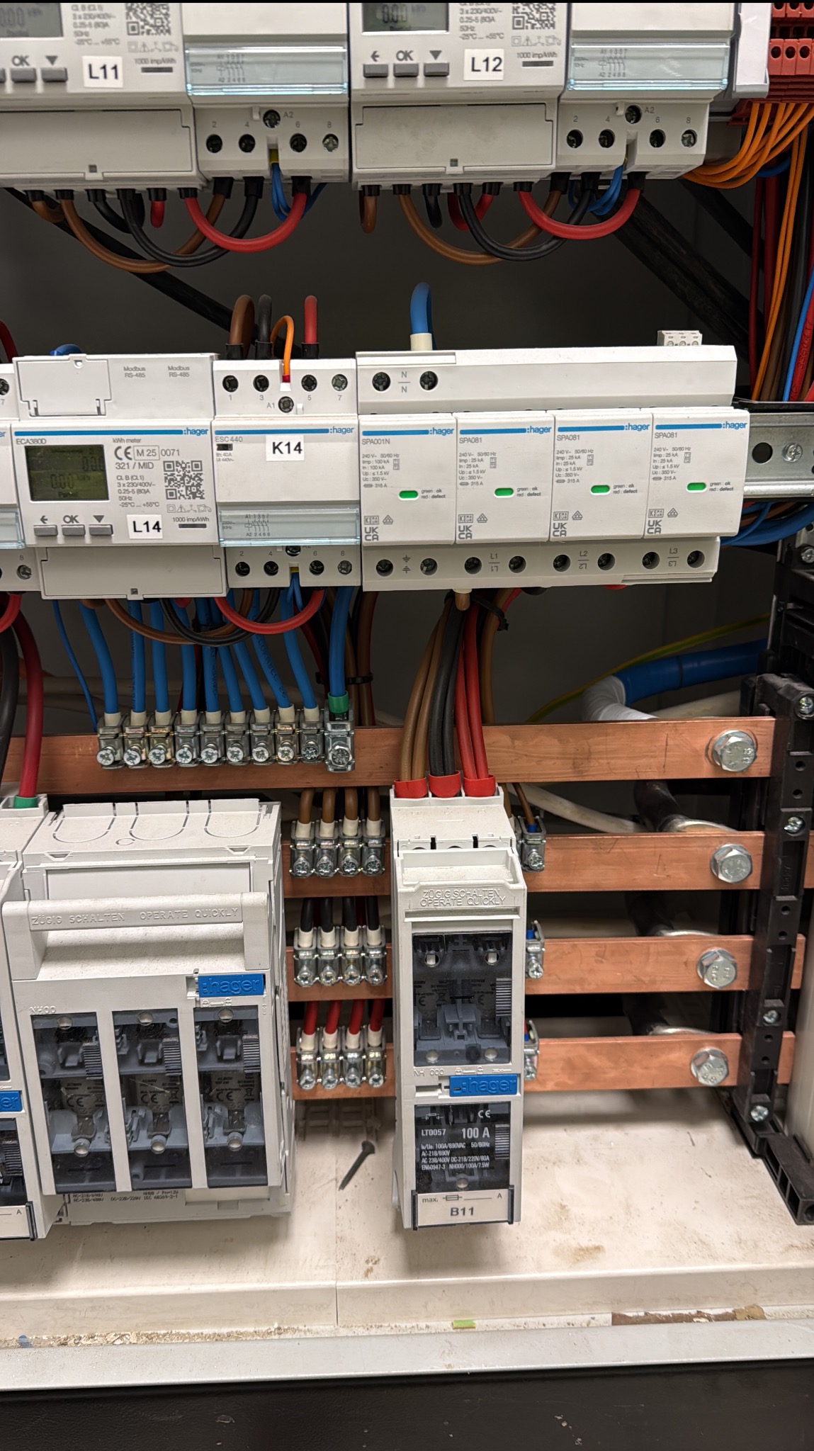 Rozdzielnica elektryczna z zabezpieczeniami, przewodami i szynami miedzianymi. Widoczne liczniki energii i bezpieczniki firmy Hager. Instalacja w kolorach niebieskim, czerwonym i brązowym.