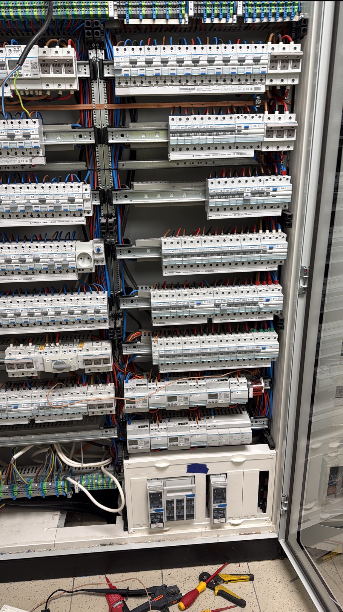 Rozdzielnica elektryczna z licznymi zabezpieczeniami, okablowaniem i narzędziami na podłodze. Widok na otwartą szafę z elementami instalacji elektrycznej.