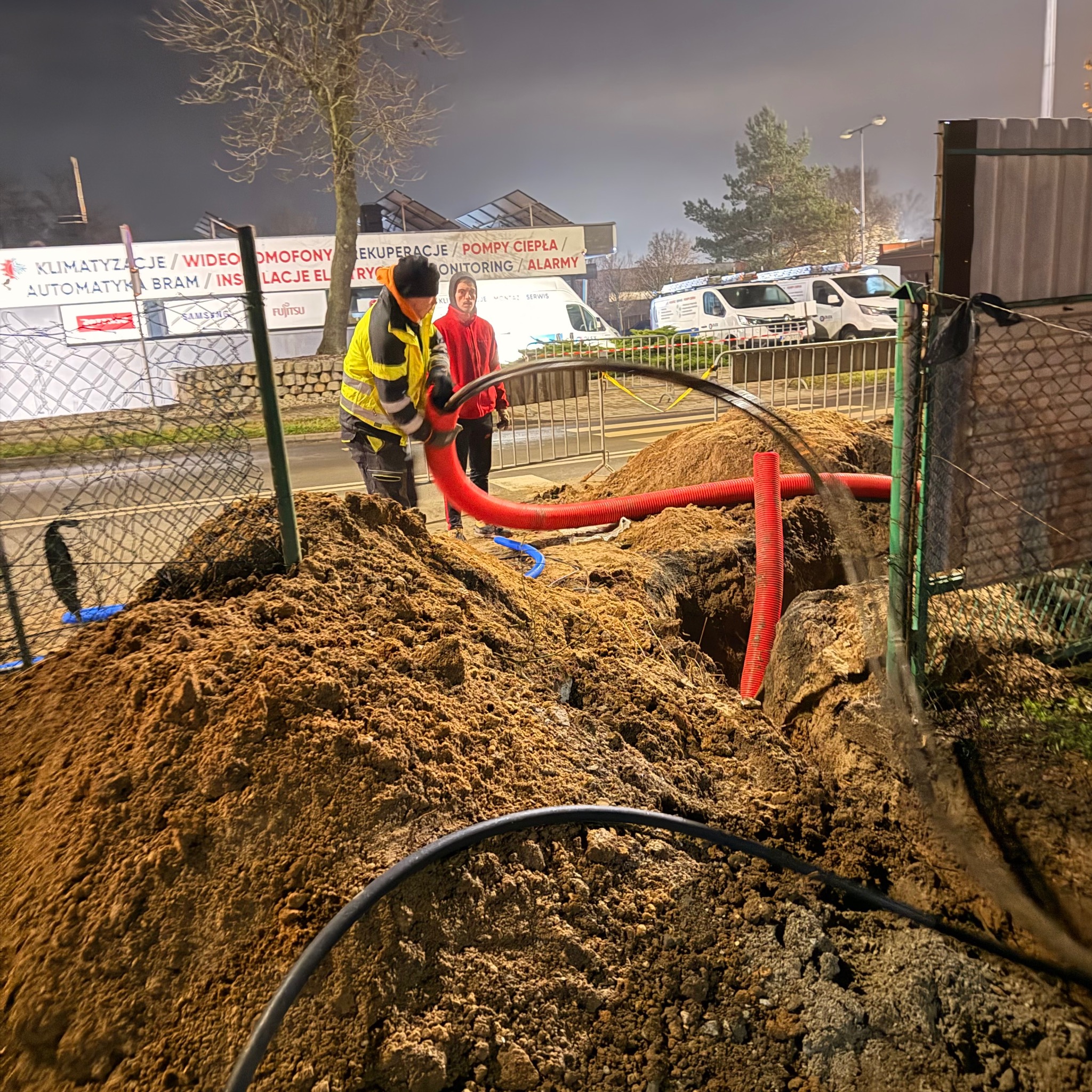 Prace ziemne i układanie rur w Złotoryi. Dwóch pracowników w odblaskowych ubraniach przy wykopie. W tle budynek z reklamą klimatyzacji i monitoringu.