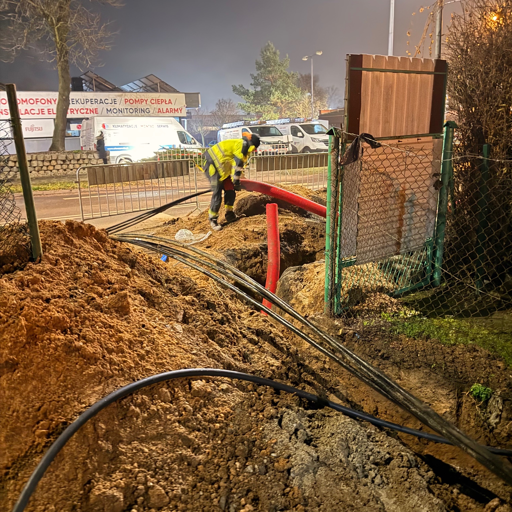 Prace ziemne przy układaniu kabli w ziemi. Widoczny pracownik w odblaskowej kamizelce i czerwone rury osłonowe. W tle budynek z reklamą instalacji elektrycznych.