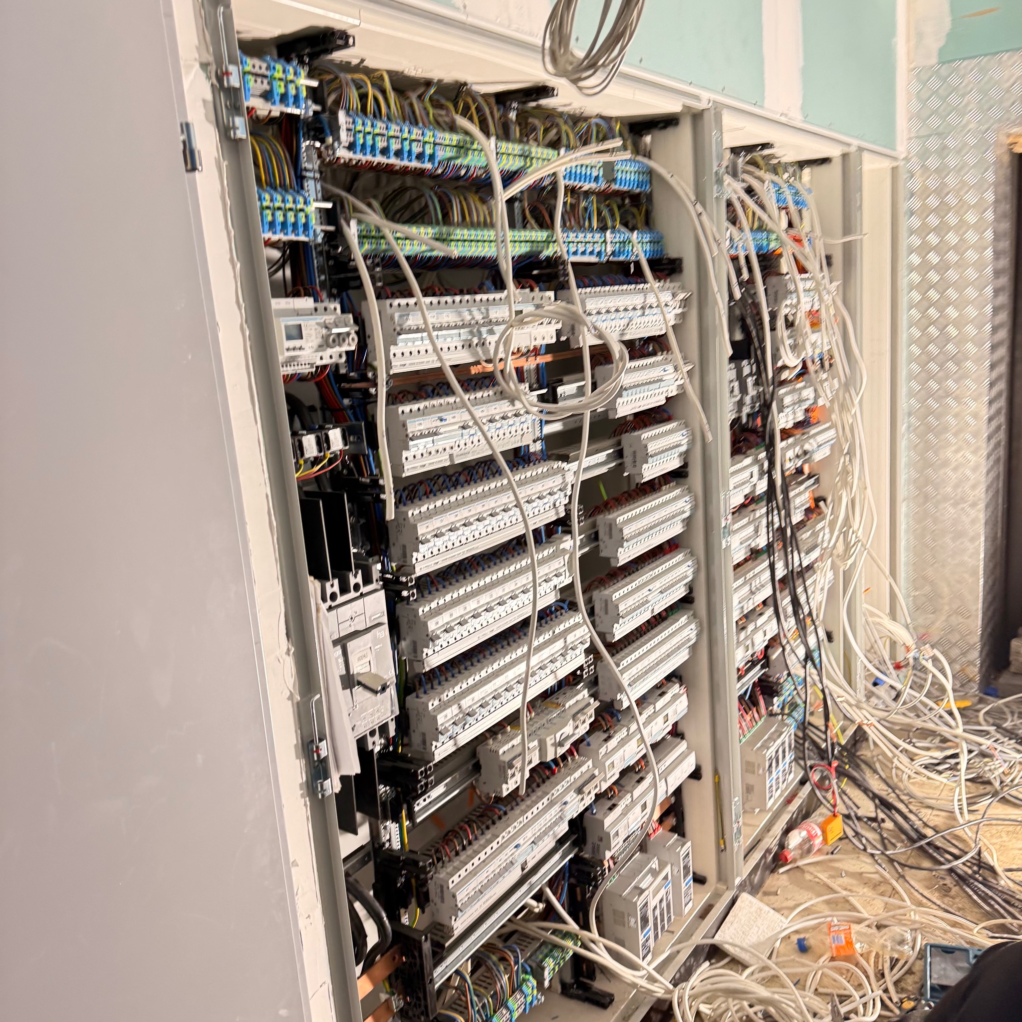 Rozdzielnica elektryczna w trakcie montażu, widoczne liczne przewody i zabezpieczenia. Częściowo zamknięta obudowa, surowy stan wykończenia wnętrza.