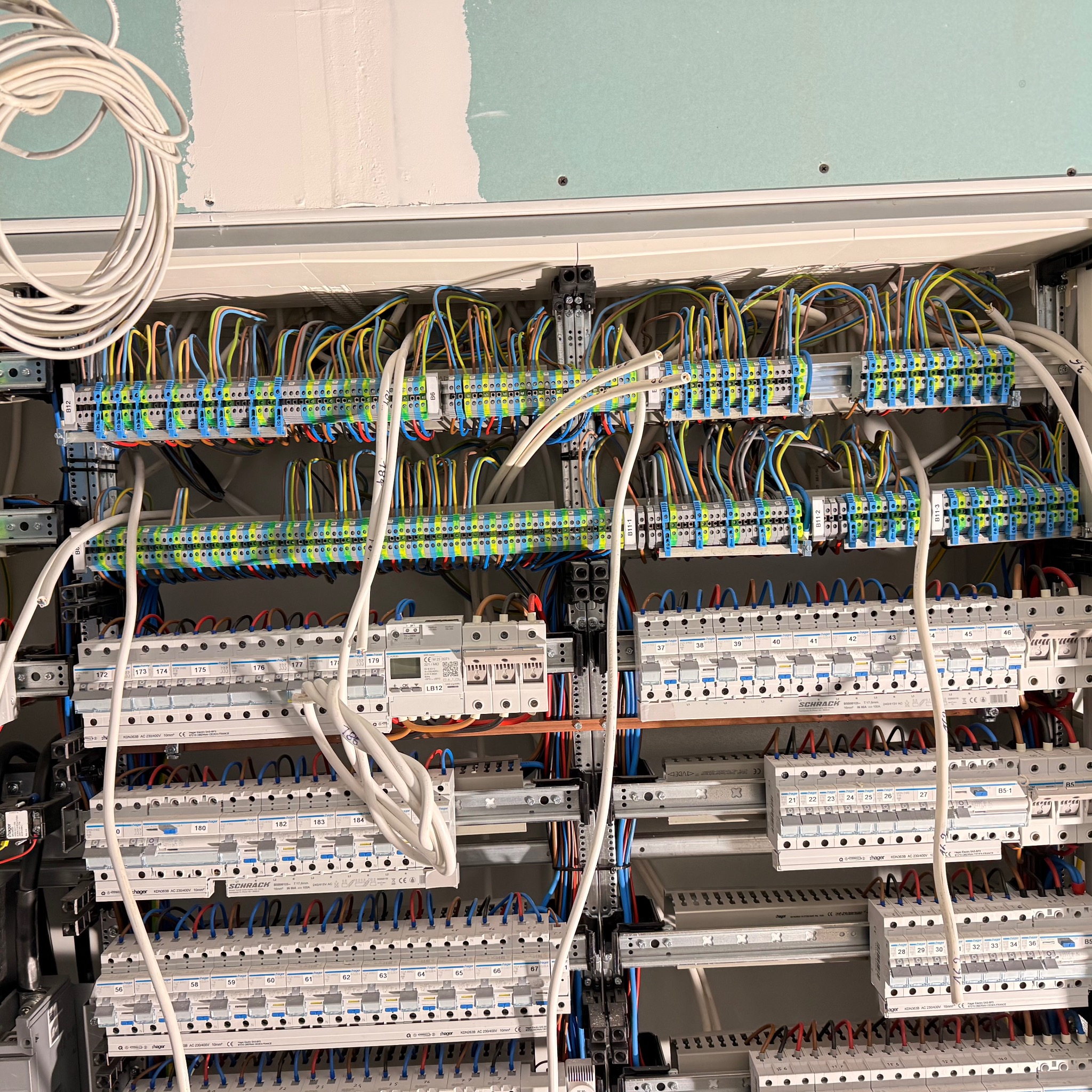 Rozdzielnia elektryczna w trakcie montażu: liczne kable w kolorowych izolacjach podłączone do listew zaciskowych i bezpieczników. Widoczne elementy marki Hager i Schrack.
