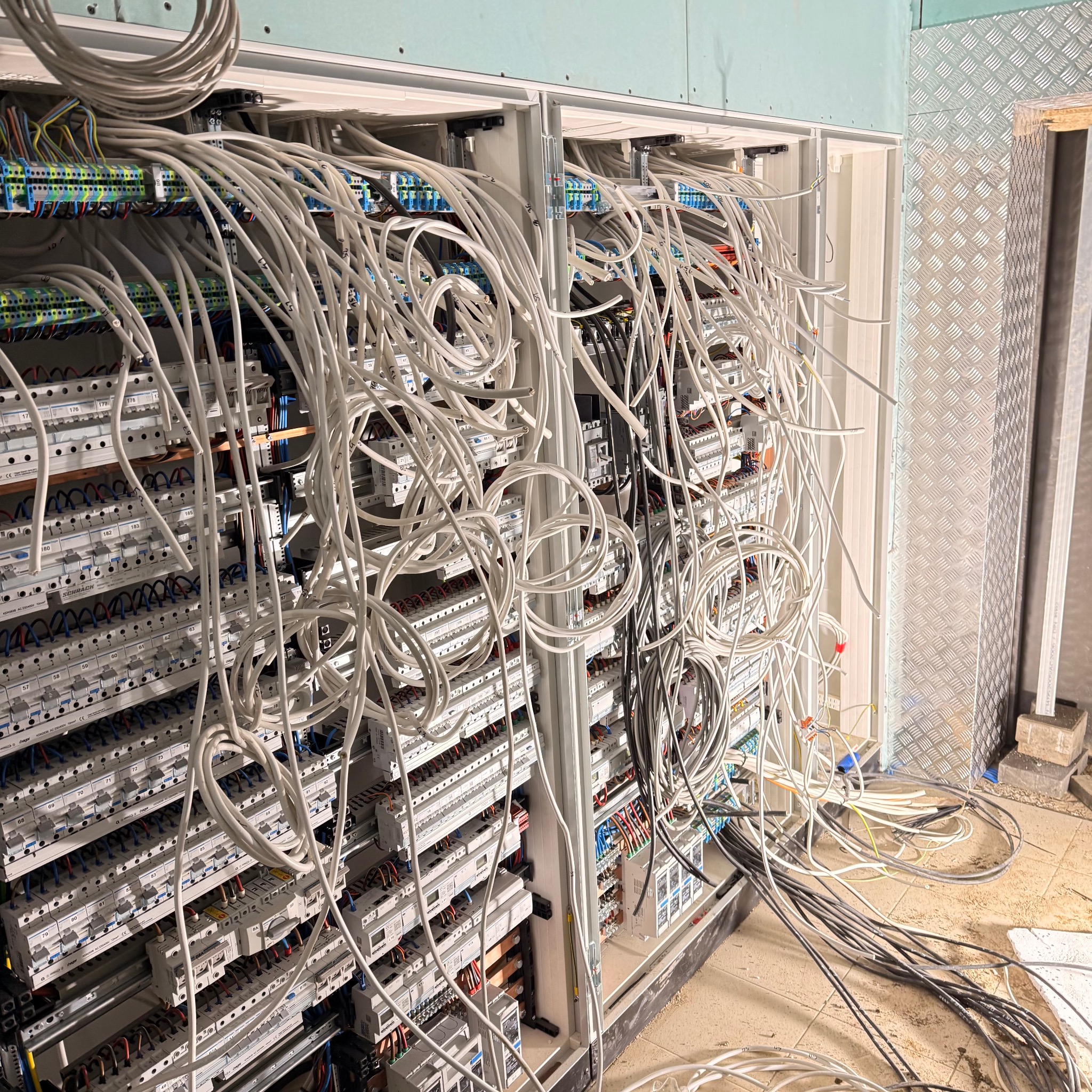 Rozbudowana szafa elektryczna z licznymi przewodami, widoczne bezpieczniki i złącza. Instalacja w trakcie prac, z elementami wykończenia wnętrz w tle.