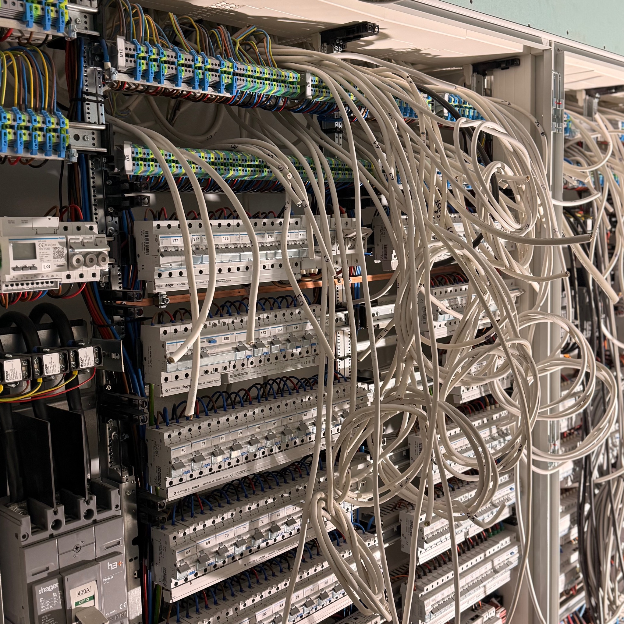 Rozbudowana skrzynka elektryczna z licznymi bezpiecznikami, przewodami i złączkami. Widoczne elementy marki Hager i Schrack. Instalacja w trakcie prac, z dużą ilością białych kabli.