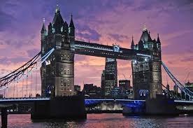Oświetlony Tower Bridge w Londynie o zmierzchu, z fioletowo-różowym niebem i odbiciem w Tamizie. Widoczne budynki w tle.